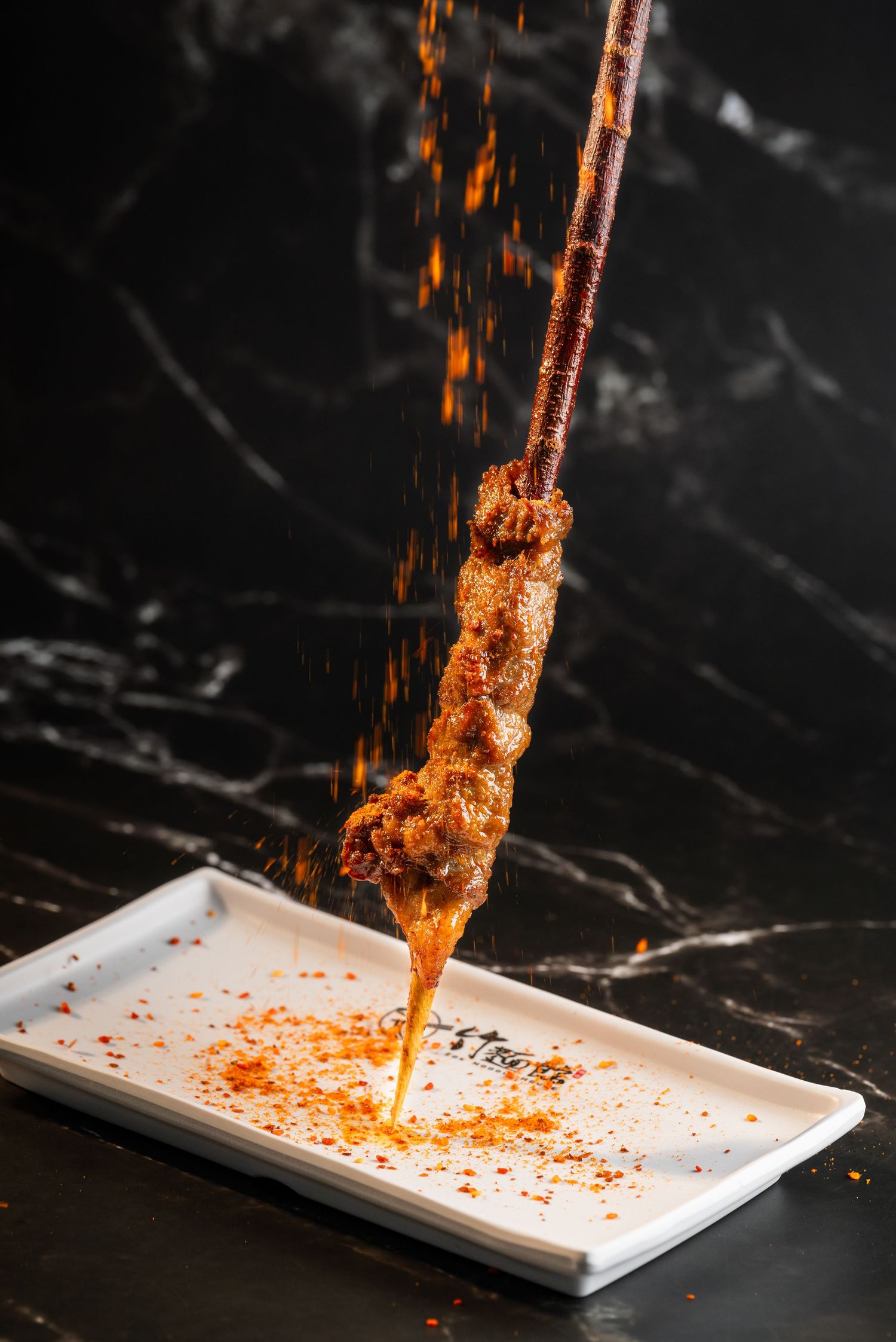 Spicy meat skewer dusted with chili powder over a white plate on a dark marble surface.