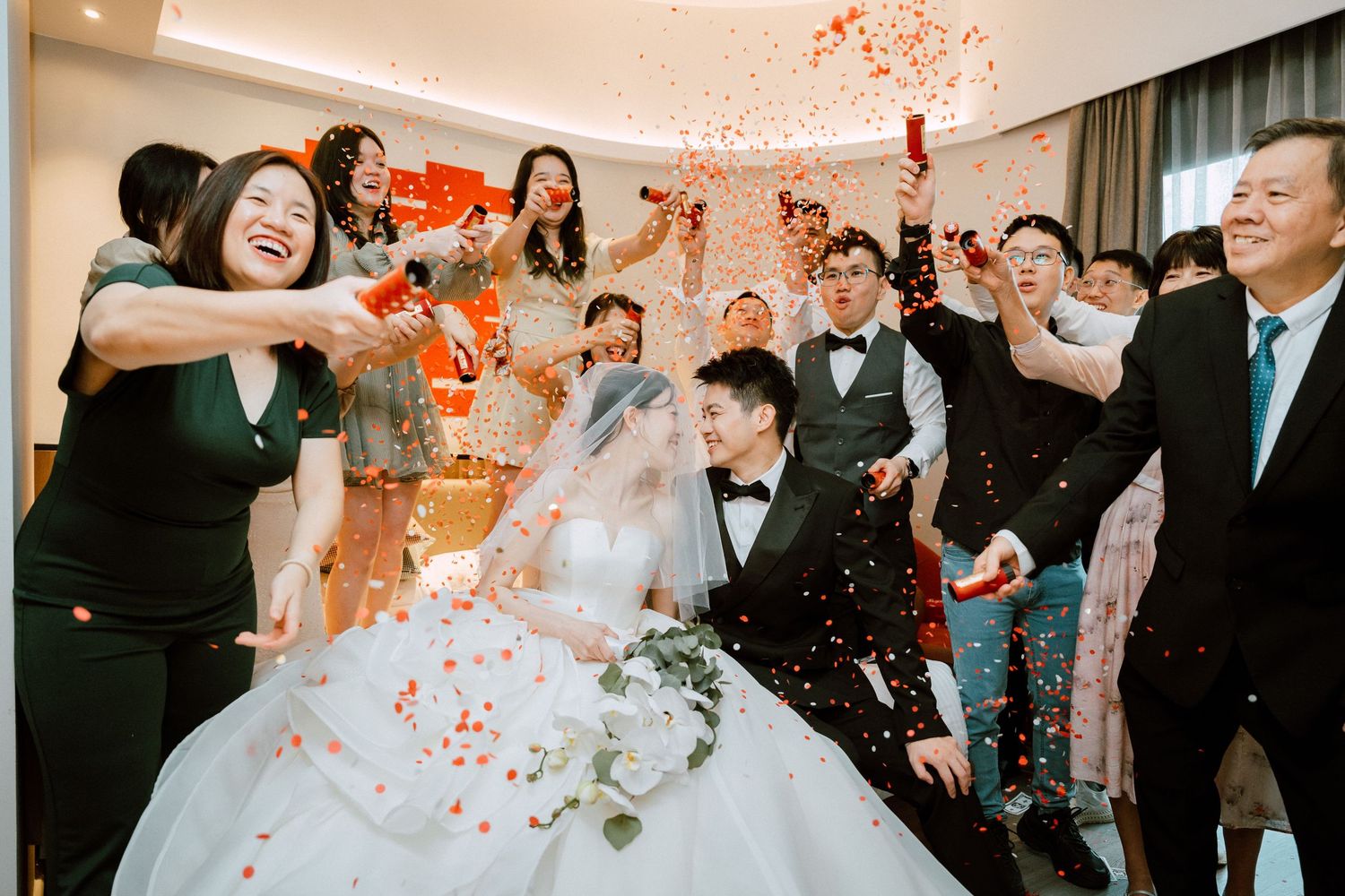 Bride and groom celebrate with guests tossing red confetti during a joyful Chinese wedding ceremony.