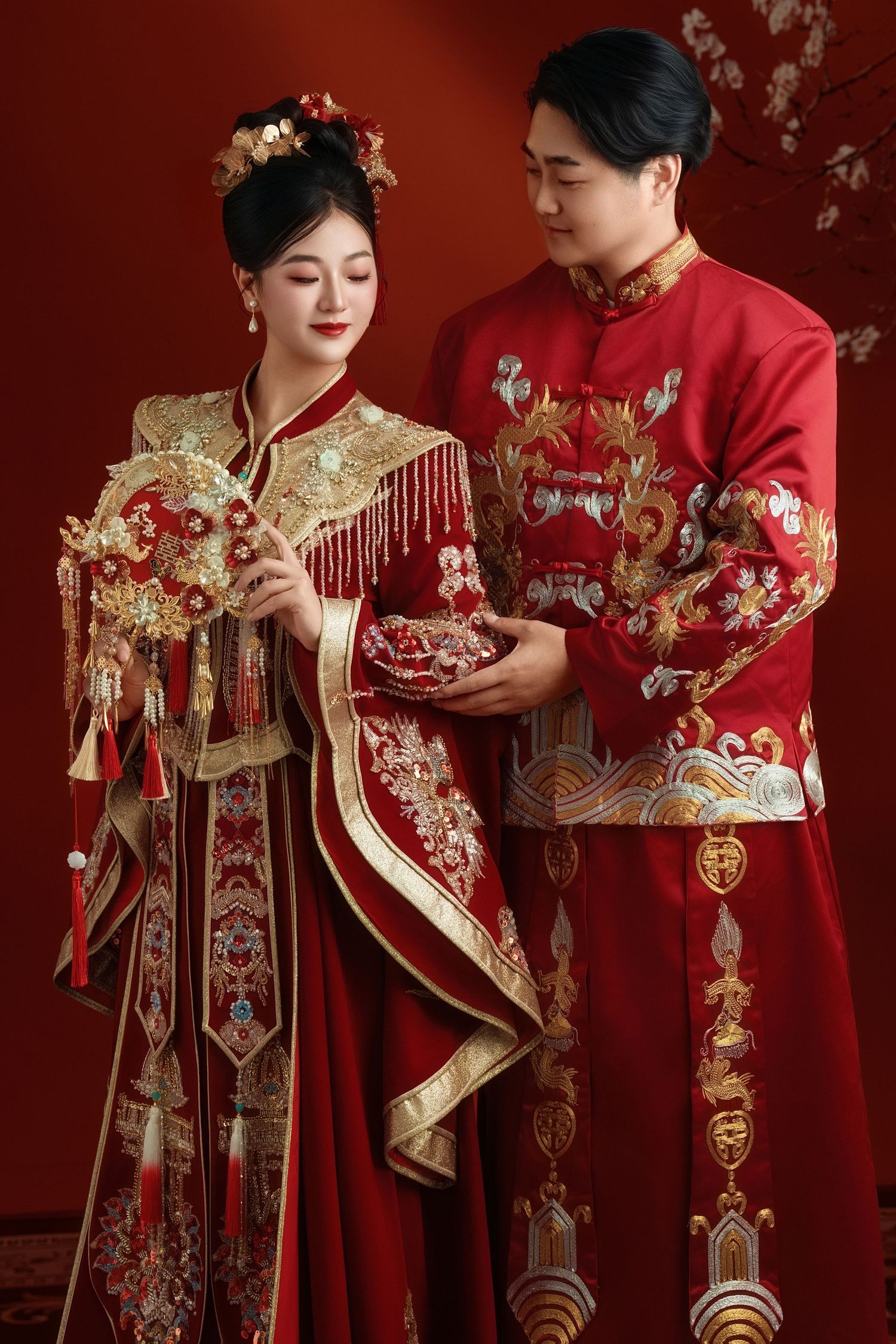 Traditional Chinese wedding couple wearing ornate red and gold formal attire with embroidered details and tassels against red backdrop.