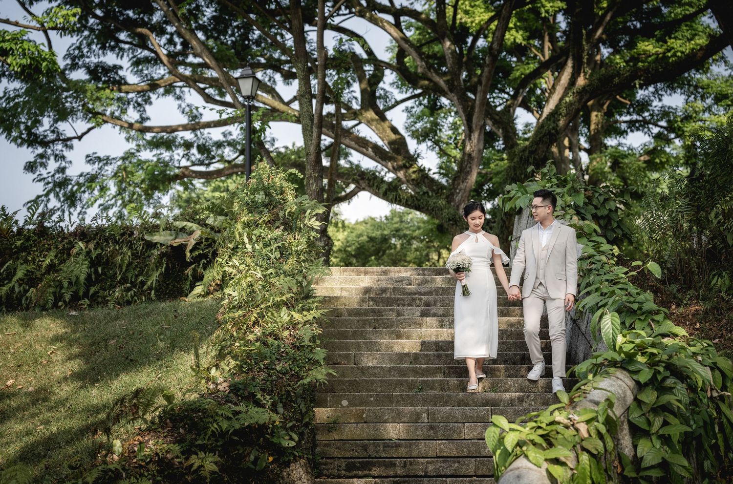 Two people stand together on stone garden steps surrounded by lush greenery on a sunny day.