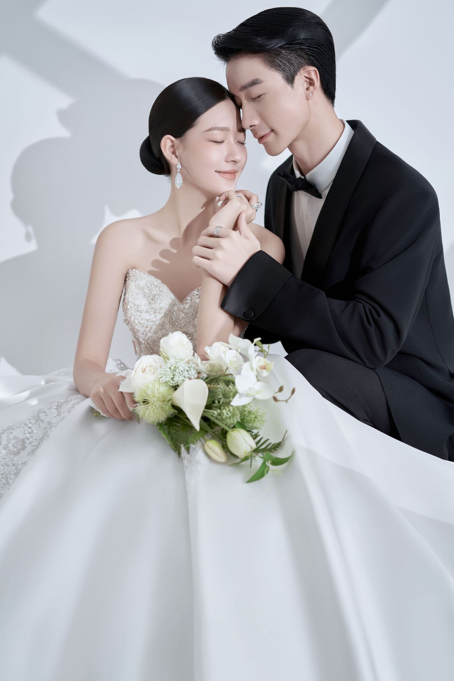 Elegant Asian couple in wedding attire, bride in white gown holding bouquet, groom in black tuxedo, romantic studio portrait.