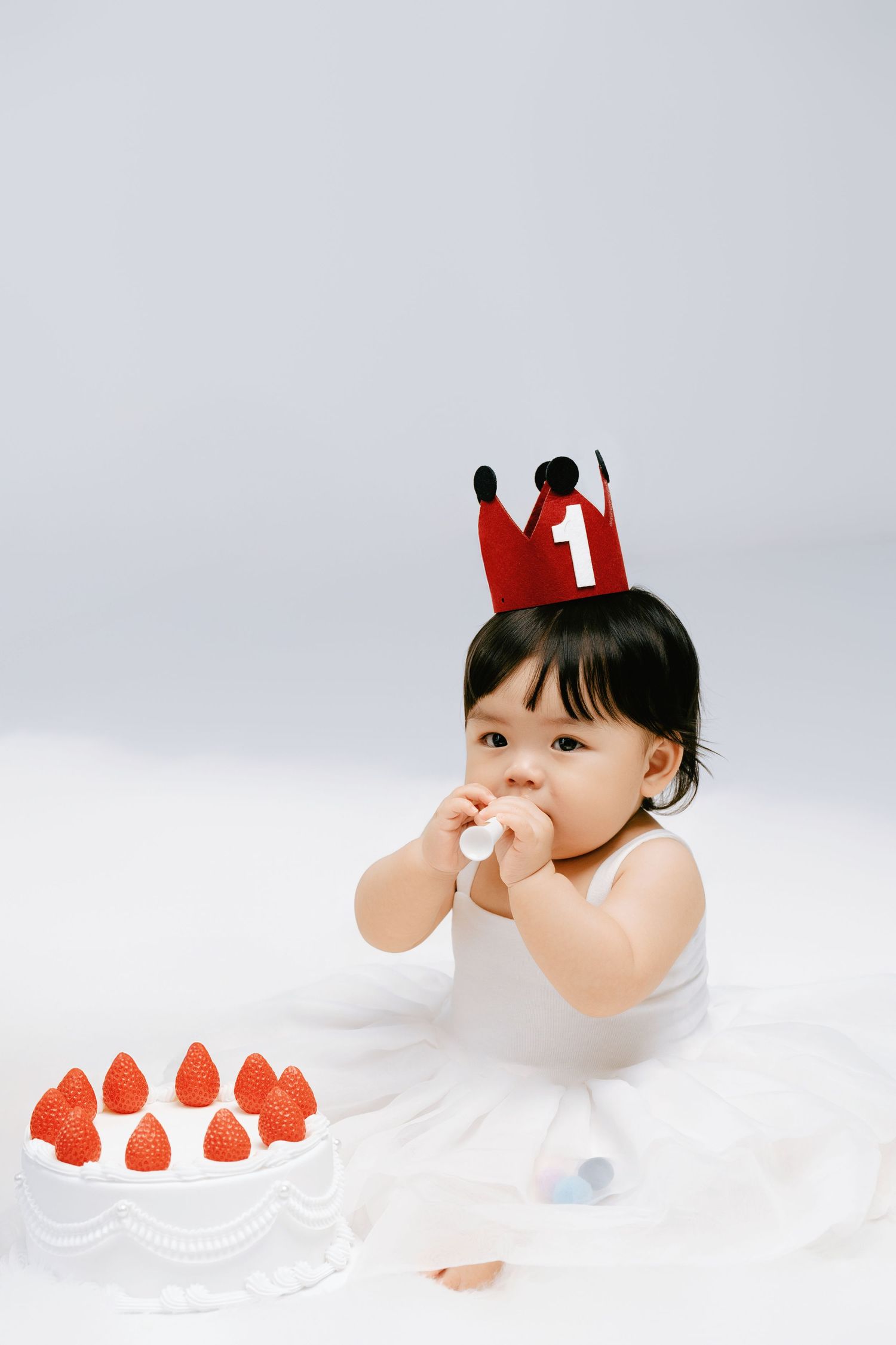 Baby girl in white dress wearing red number 1 crown sits beside strawberry birthday cake celebrating her first birthday.