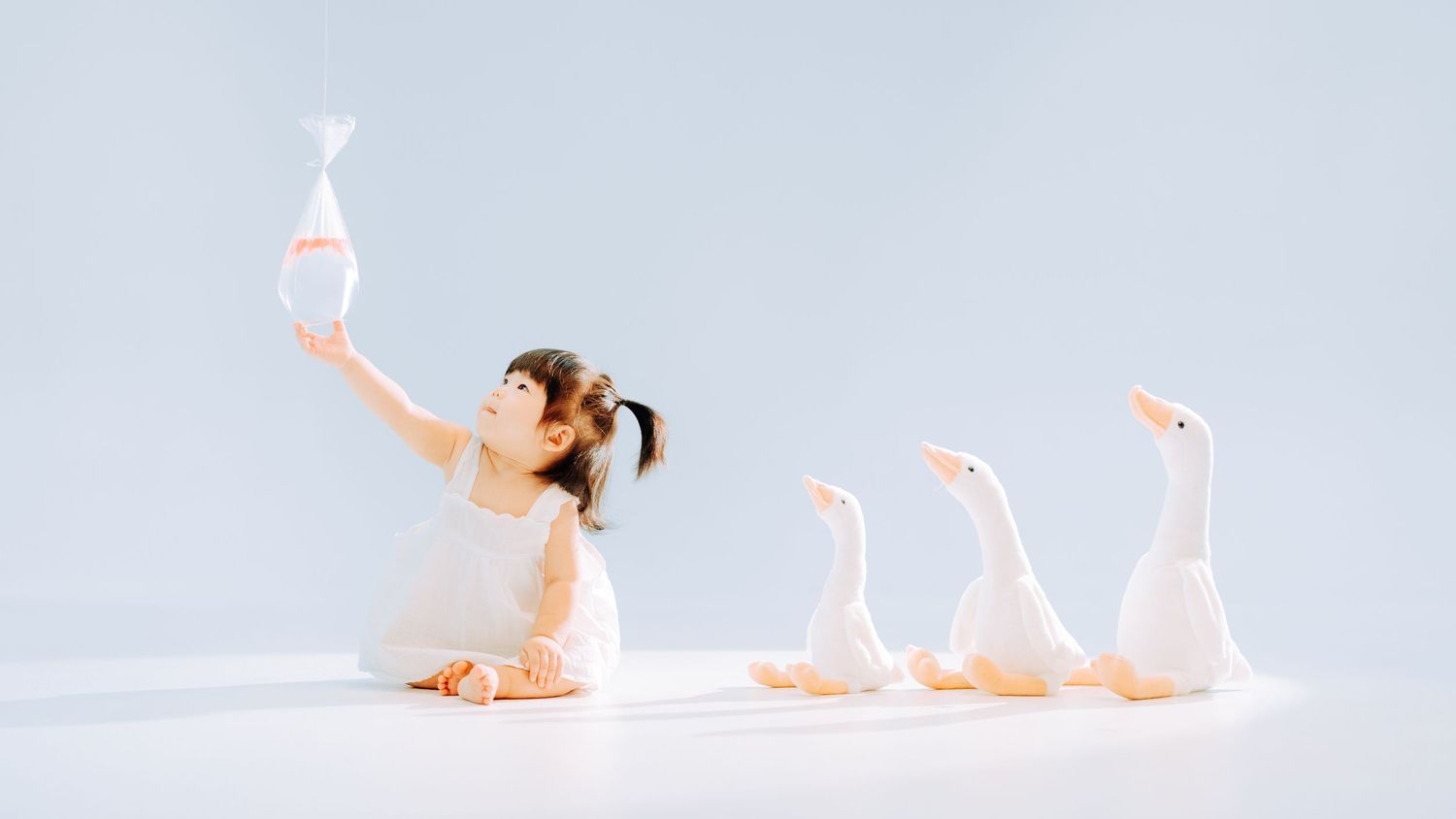 A young girl sits on a white floor, reaching up with a toy fish, surrounded by three white duck figurines.