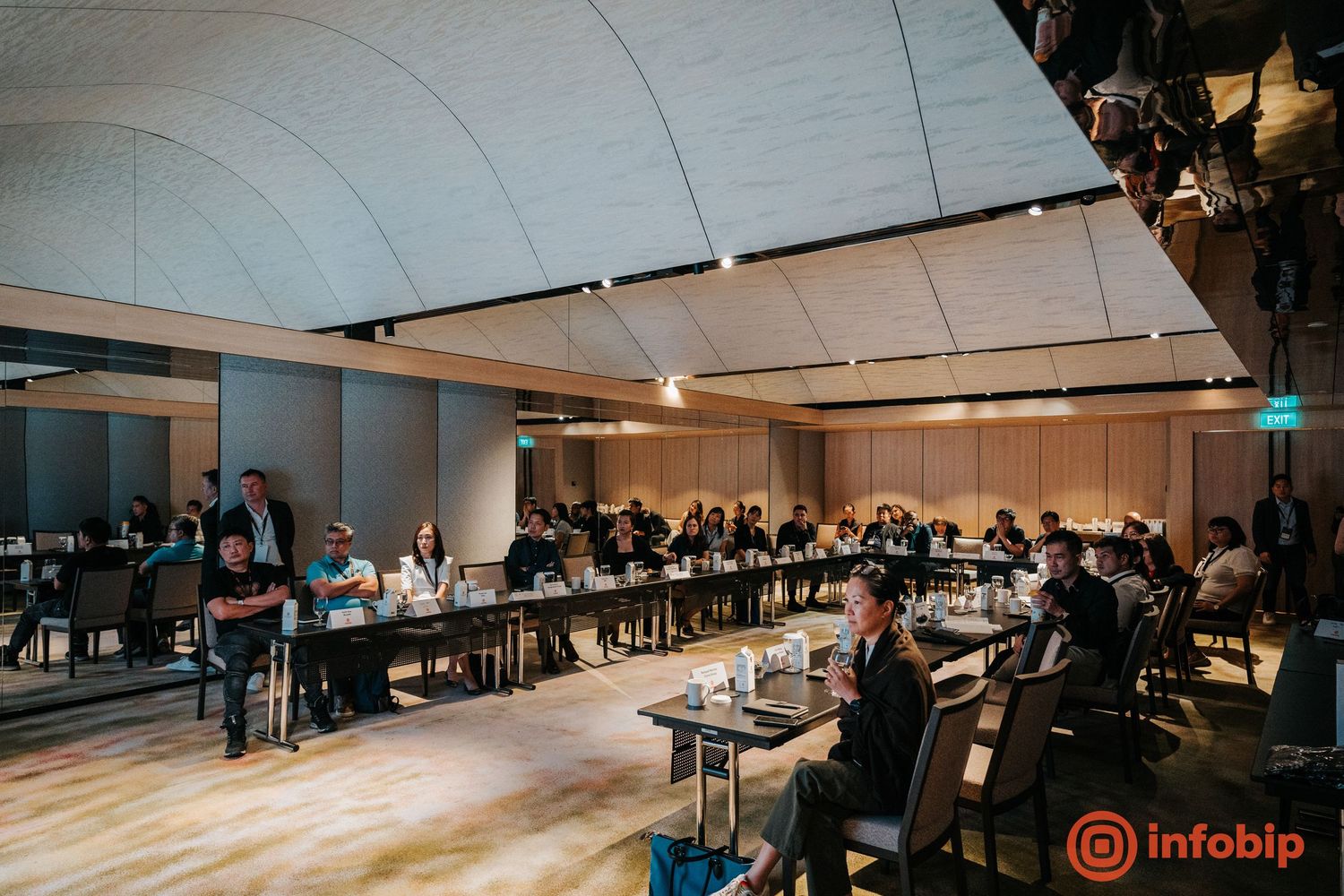 Business conference attendees seated at long tables in a modern event hall with curved ceiling panels.