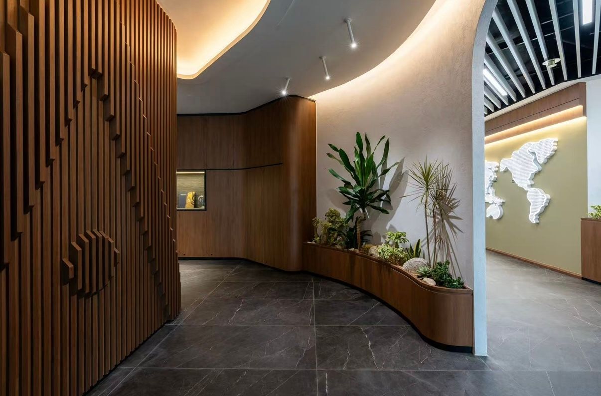 Modern office lobby with curved wooden walls, lush tropical plants, and elegant dark tile flooring under warm lighting.