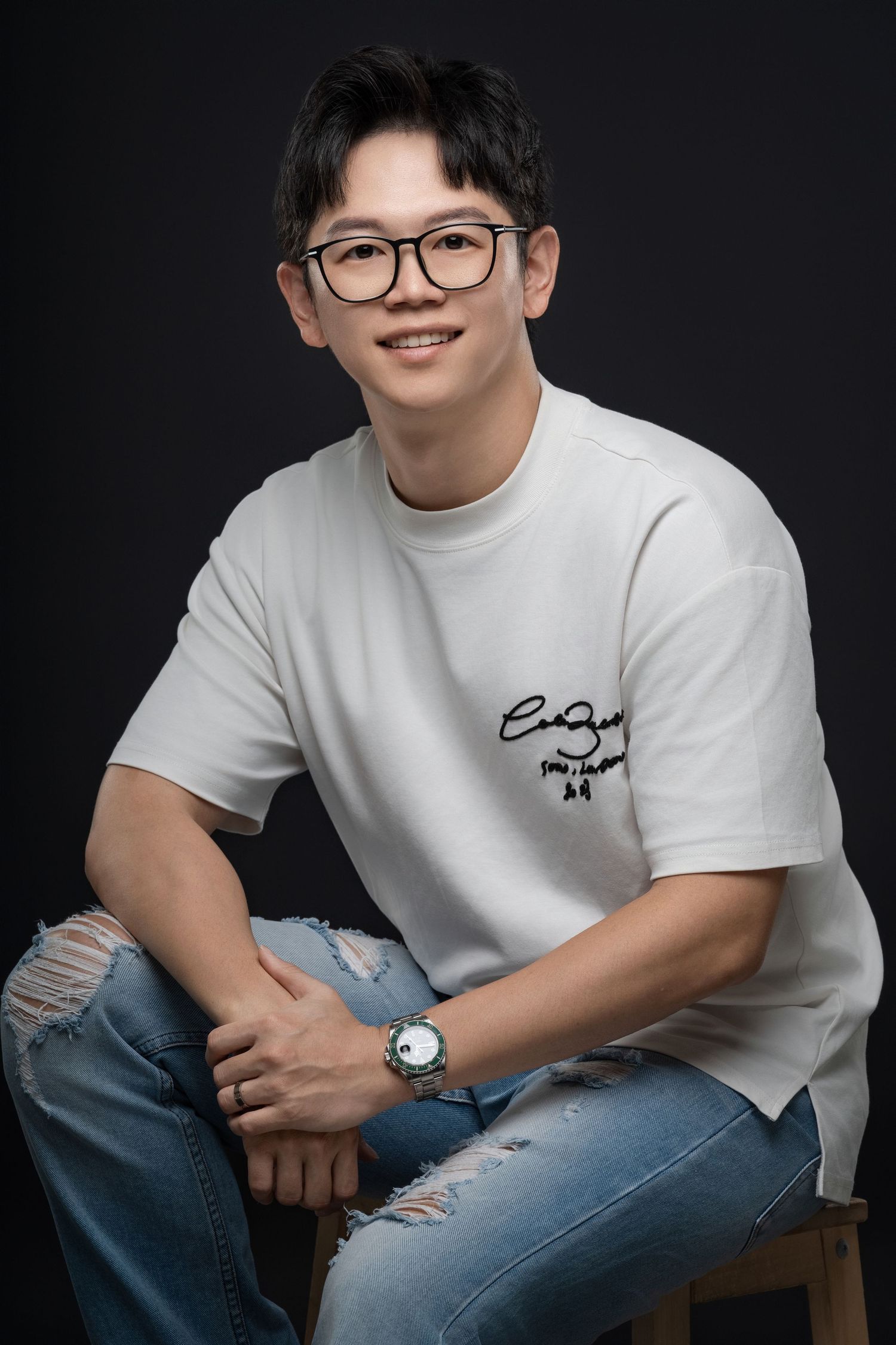 Young Asian man smiling, wearing glasses, white graphic tee, ripped jeans, and a watch, posed on a stool against dark background.