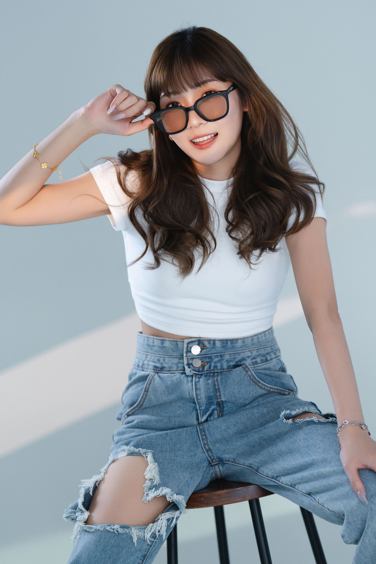 Young Asian woman in white crop top and ripped jeans sitting on stool, adjusting oversized sunglasses, smiling.
