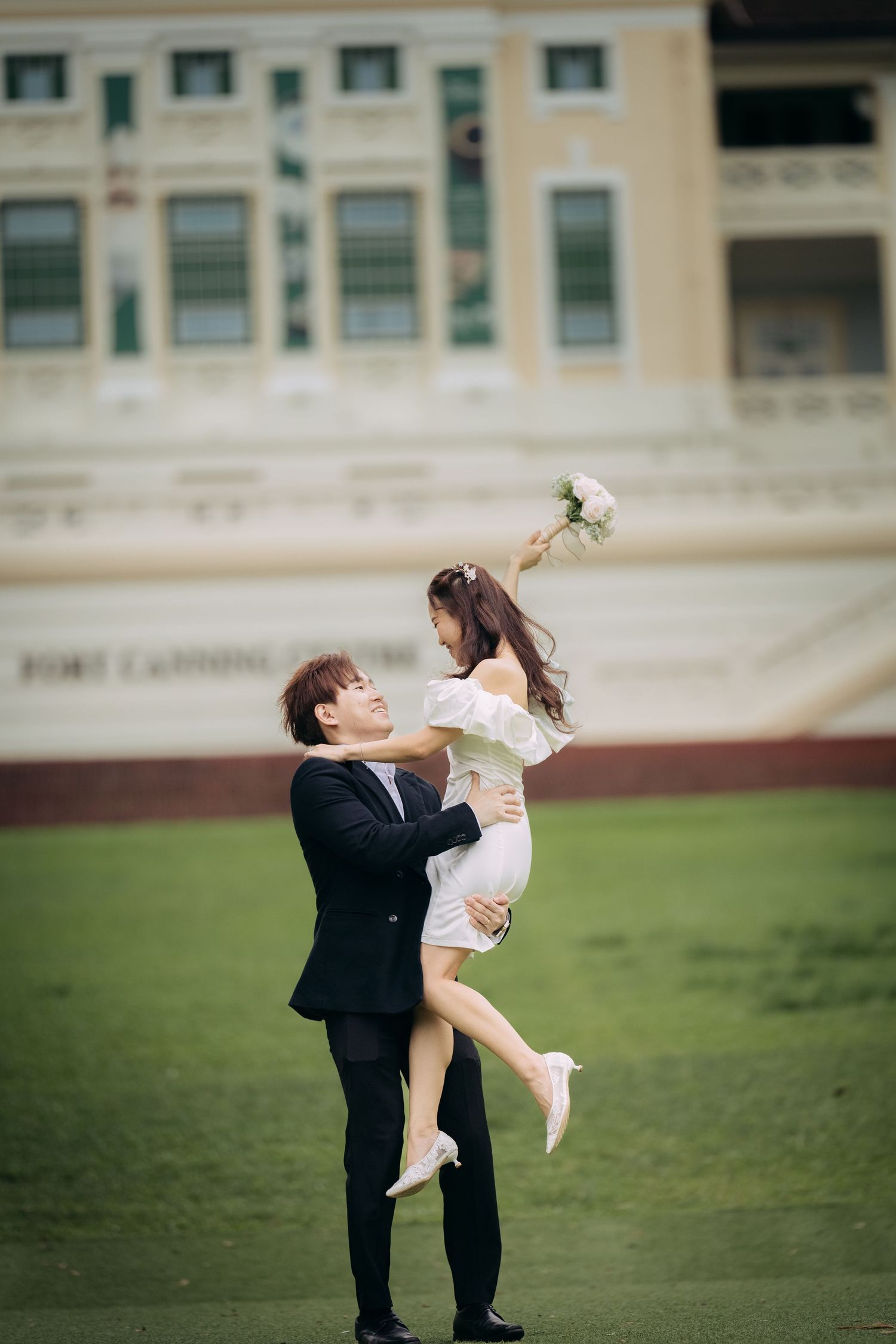 Groom lifting bride in white dress outdoors on green field, holding bouquet, with historic building backdrop.