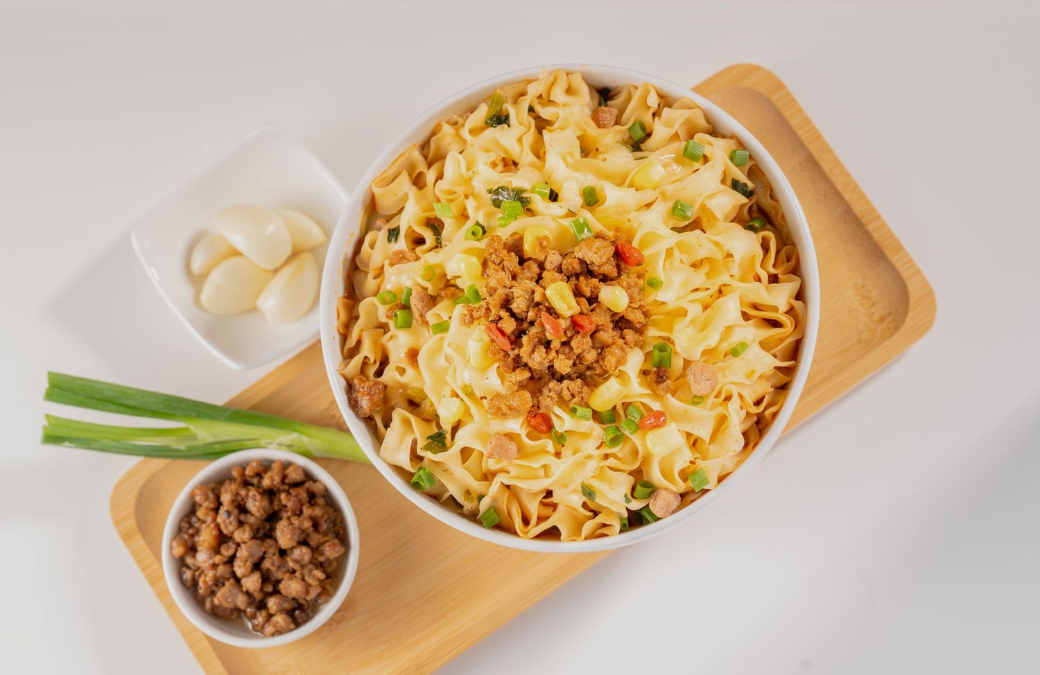 Bowl of egg noodles topped with minced meat and green onions, served on a wooden tray with garlic cloves.