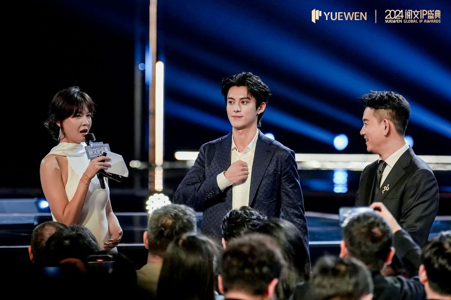 Three elegantly dressed presenters on a glamorous awards show stage with YUEWEN branding visible in the background.