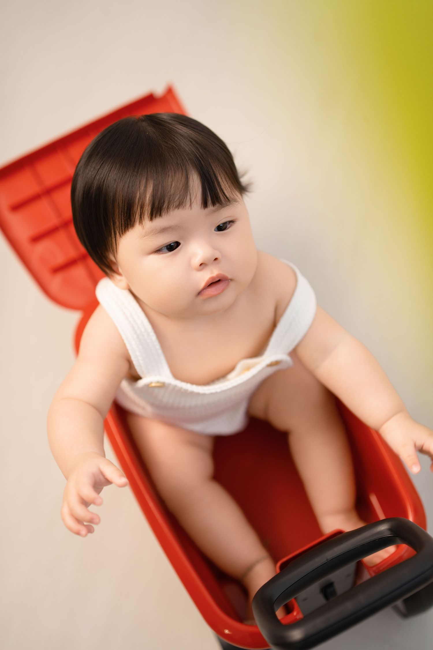 Small child wearing white top sits in red plastic chair while looking down with curiosity.