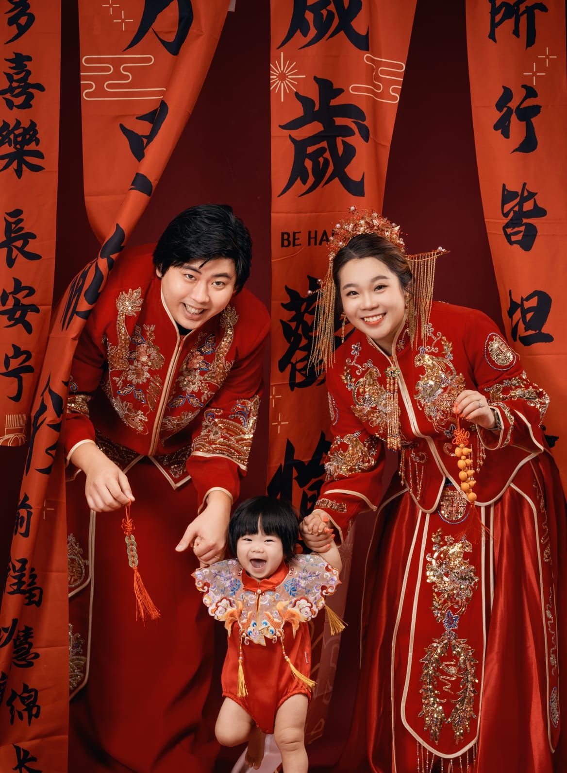 Traditional Chinese New Year family photo with people wearing red and gold qipao dresses against a backdrop of calligraphy banners.