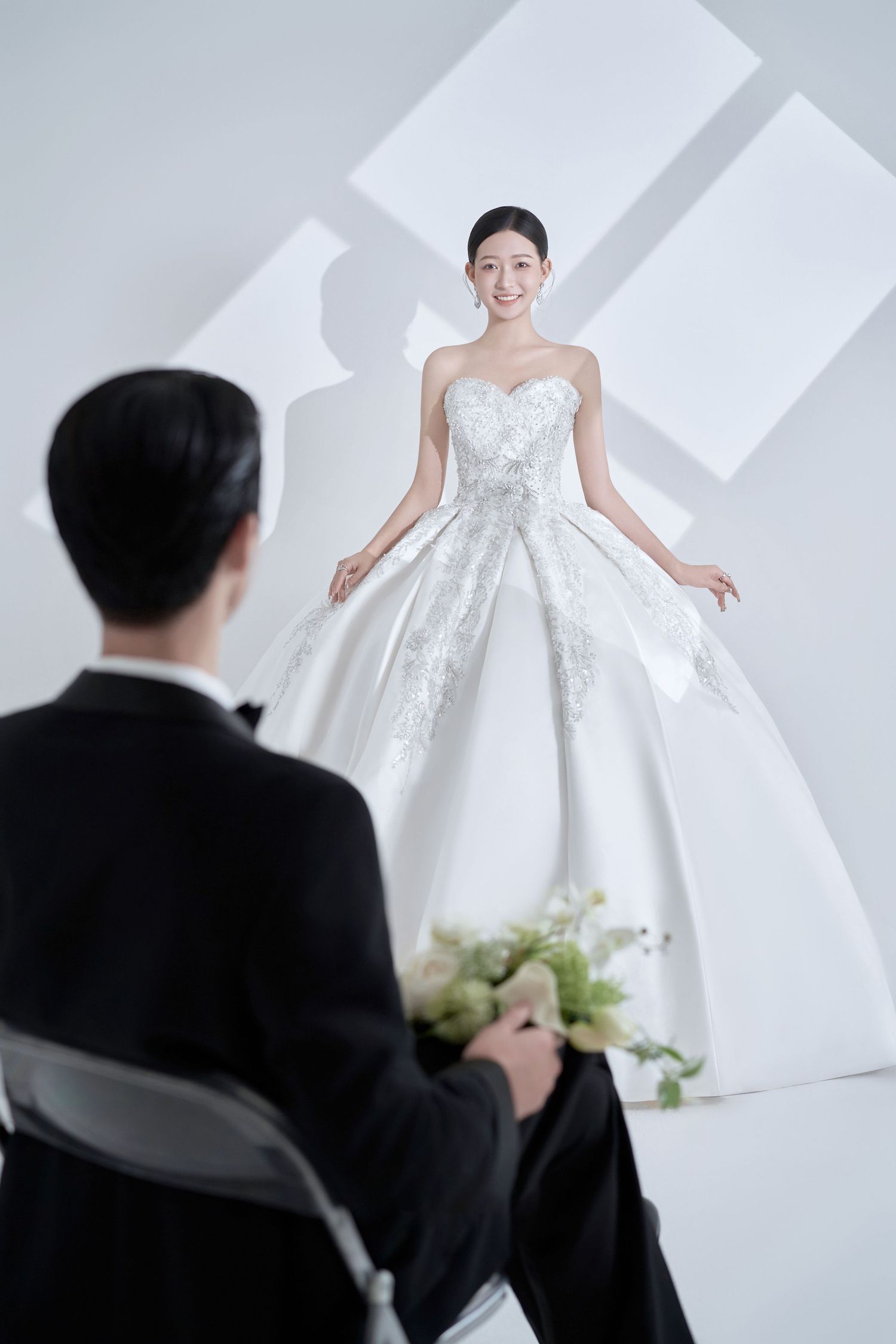 A bride in a white ballgown smiles at a groom in a tuxedo holding a bouquet during a wedding photo shoot.