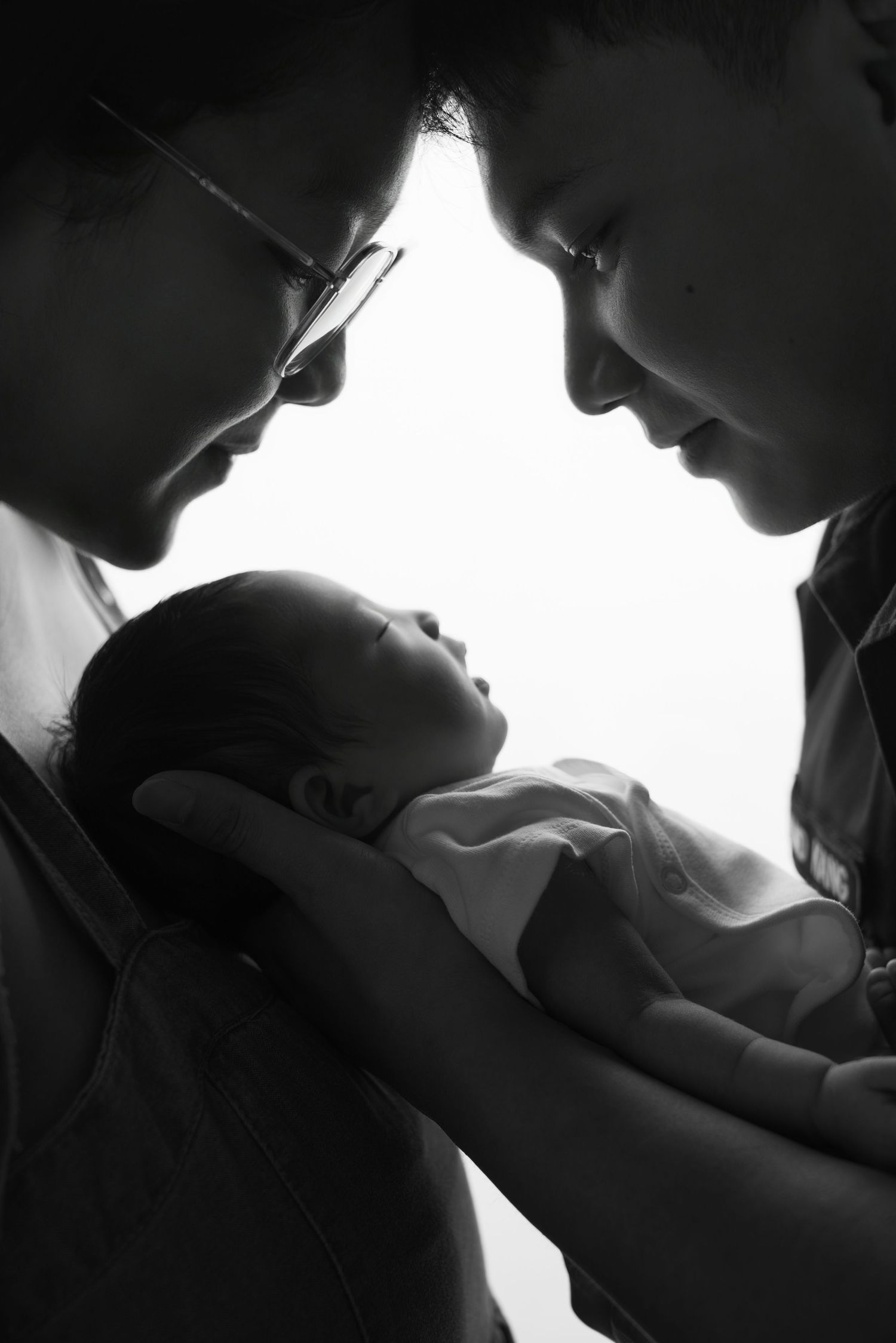 Black and white photo of two parents lovingly cradling their newborn baby, heads bowed together in tender moment.