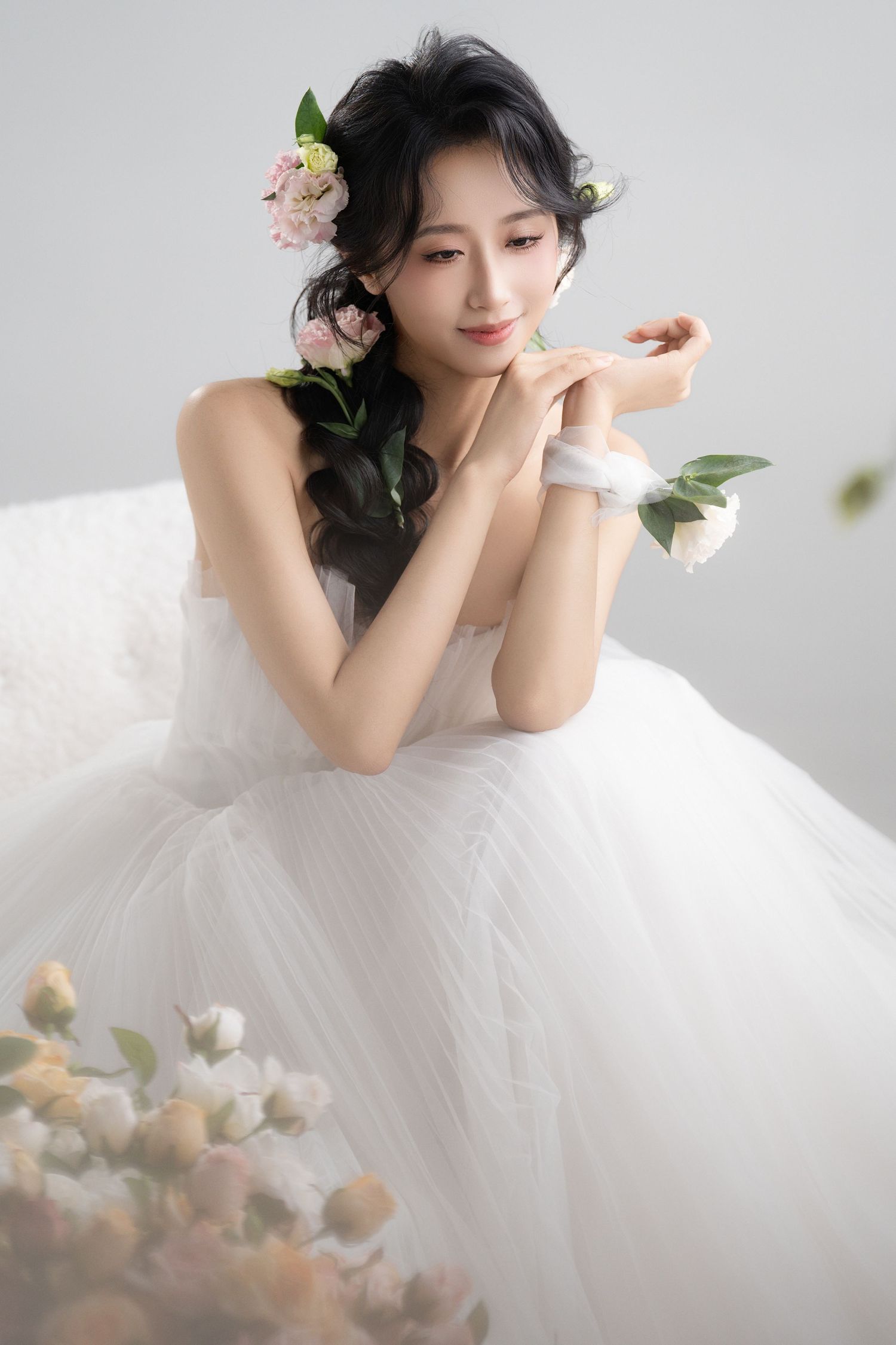 Young Asian woman in white tulle wedding dress with flowers in hair, posing gracefully in a soft studio setting.