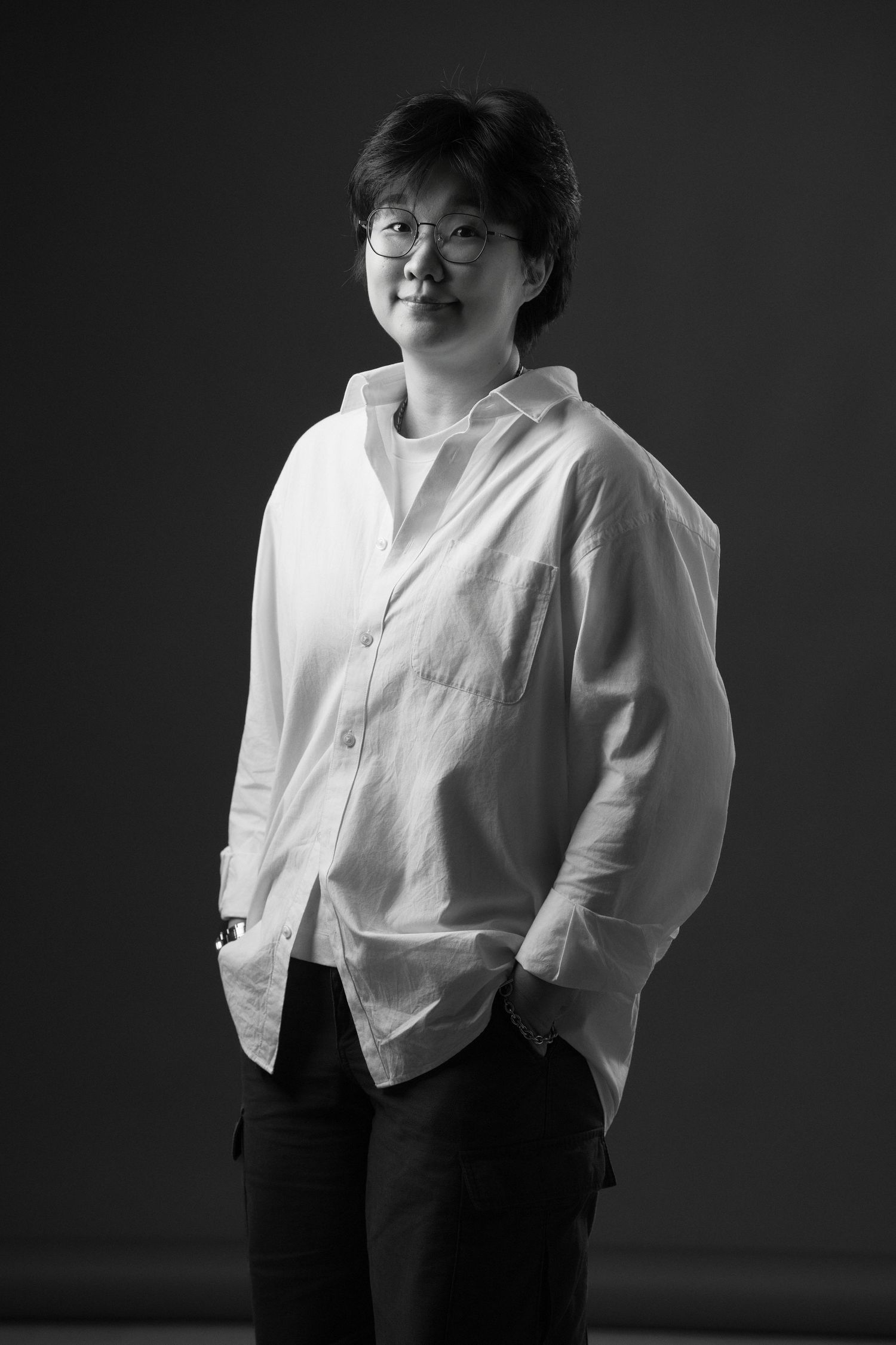 Black and white portrait of a person wearing glasses and a white shirt, hands in pockets, smiling softly.