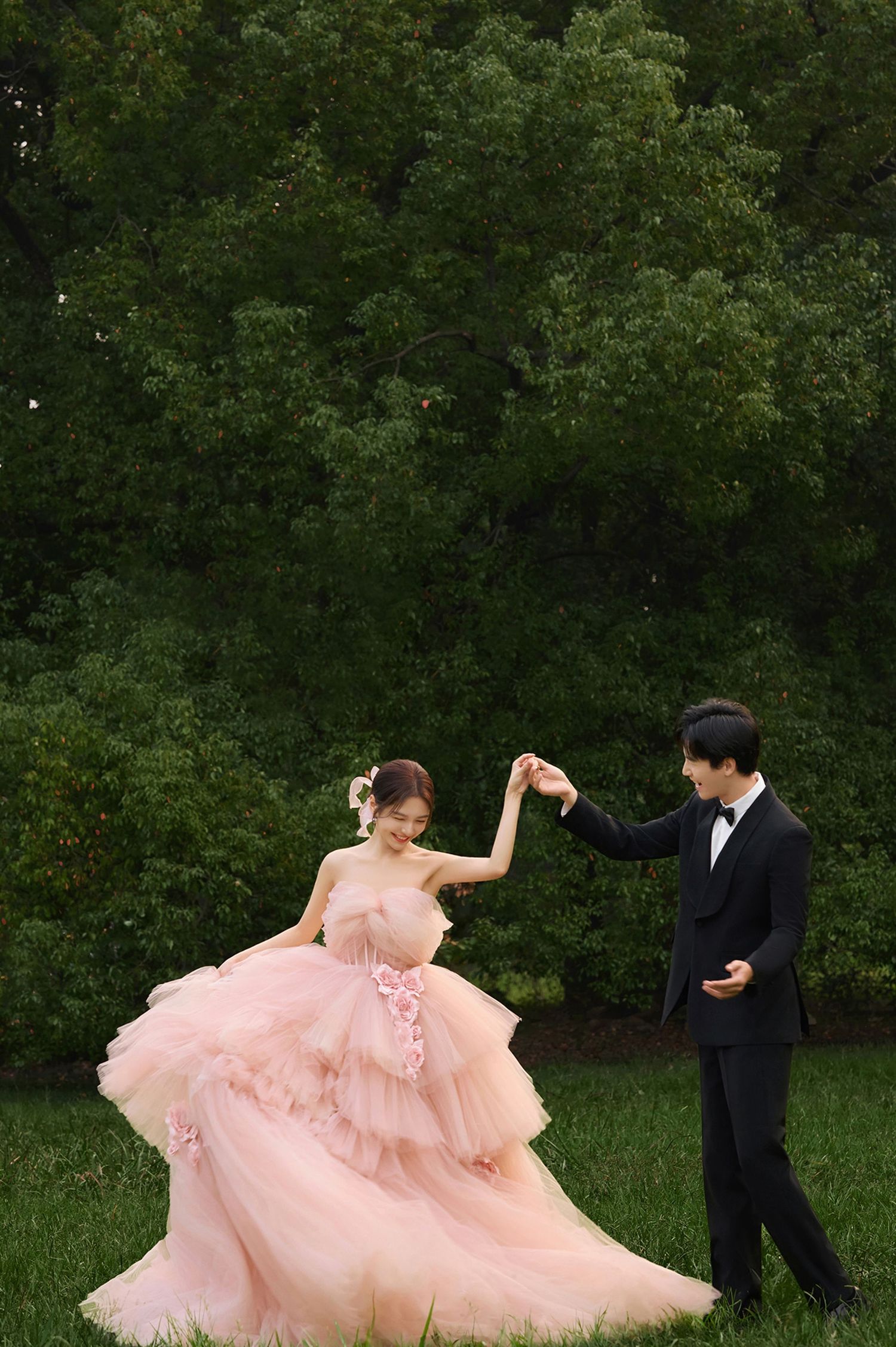 A couple in formal attire dance outdoors, with the bride wearing a flowing pink tulle gown against a backdrop of green trees.