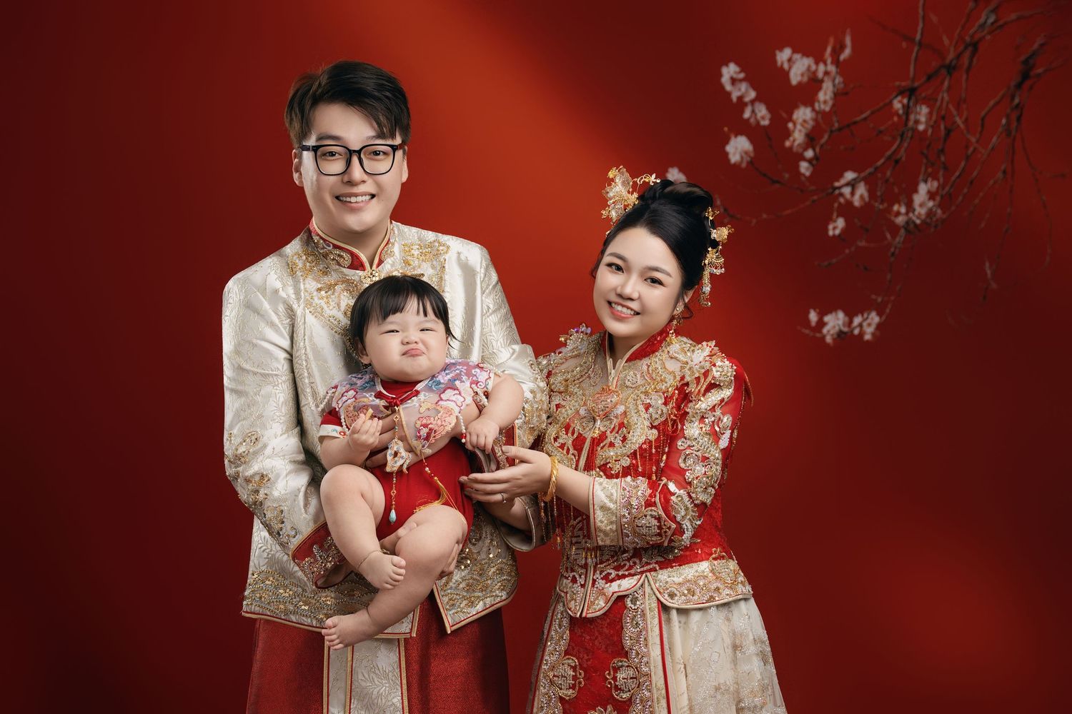 Chinese family in traditional red and gold qipao outfits posing with baby for festive portrait against red background.