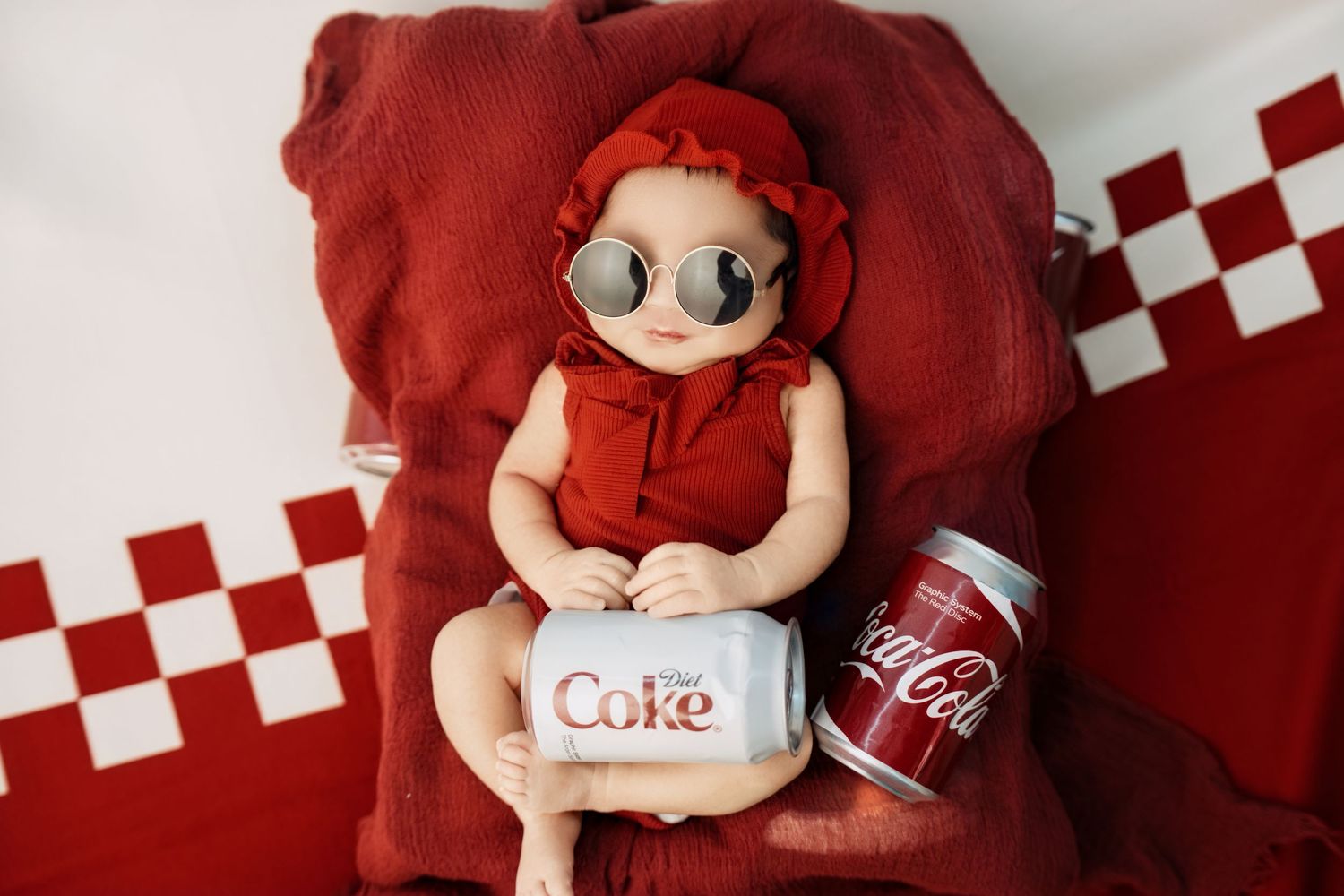 Baby in red outfit and sunglasses poses with Diet Coke and Coca-Cola cans on a red cushioned chair.