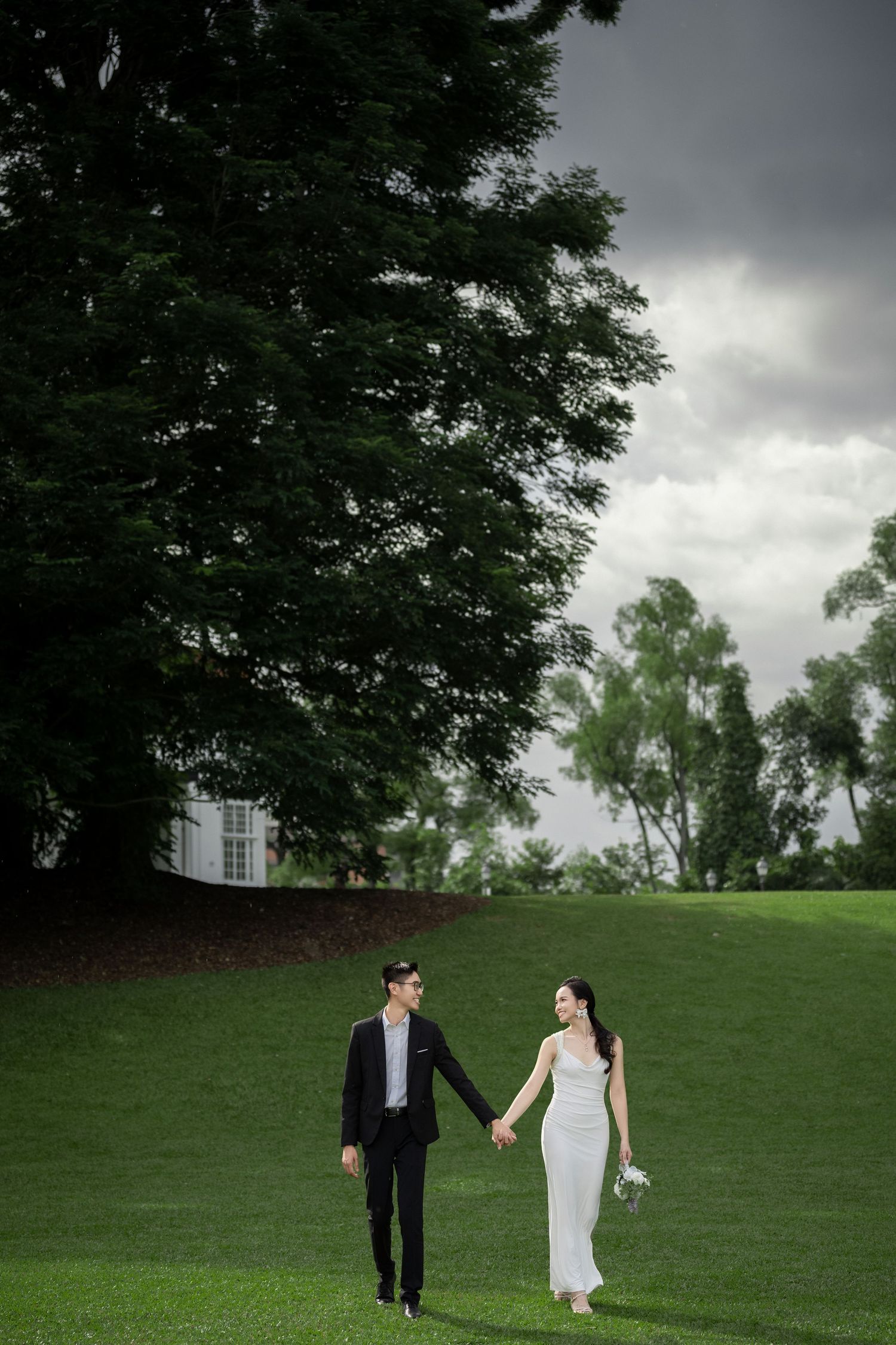 A wedding couple holds hands while walking on a lush green hillside under moody storm clouds approaching.