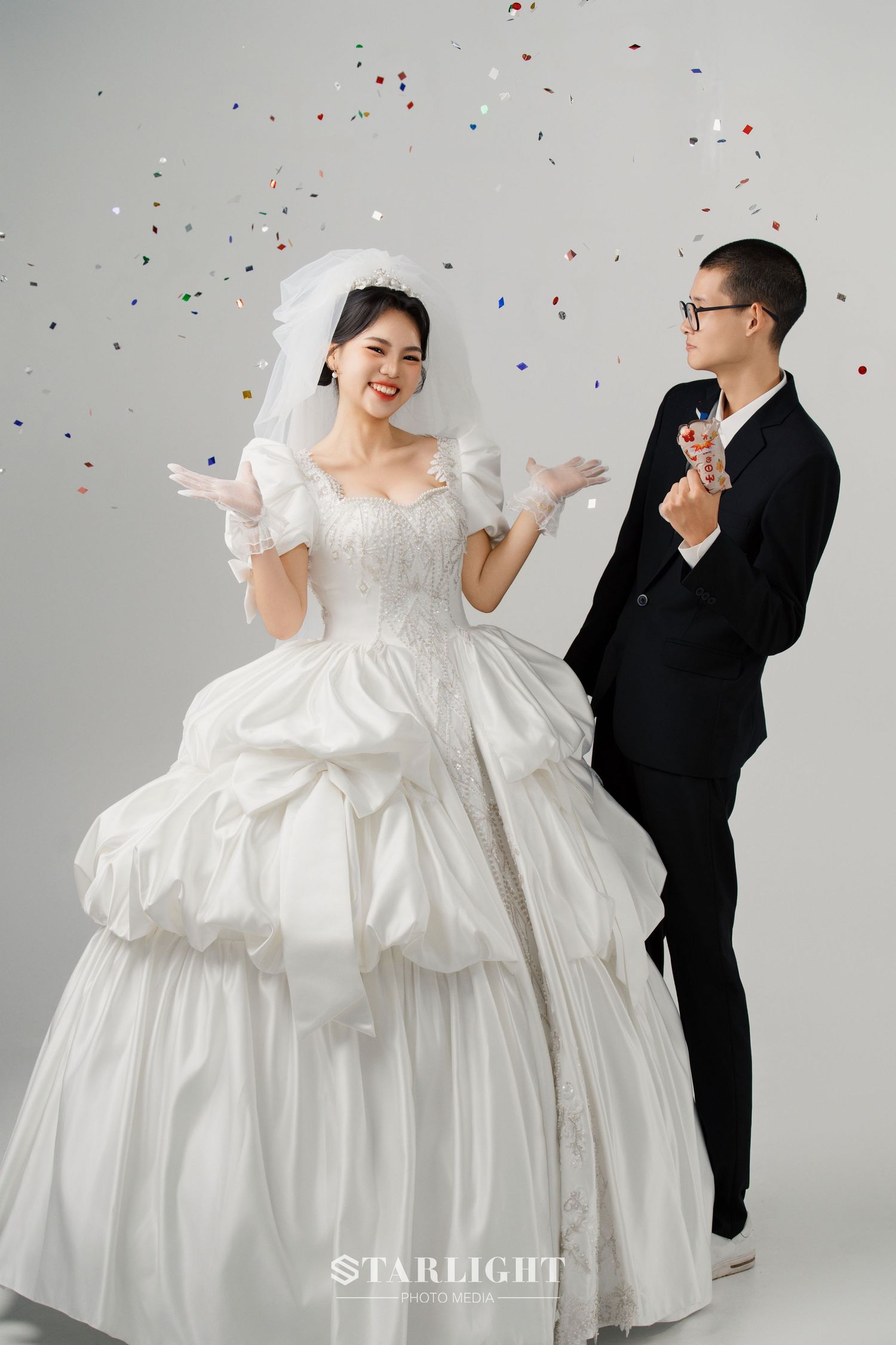 A couple poses playfully amid falling confetti wearing a white wedding gown and black suit in a series of studio photos.
