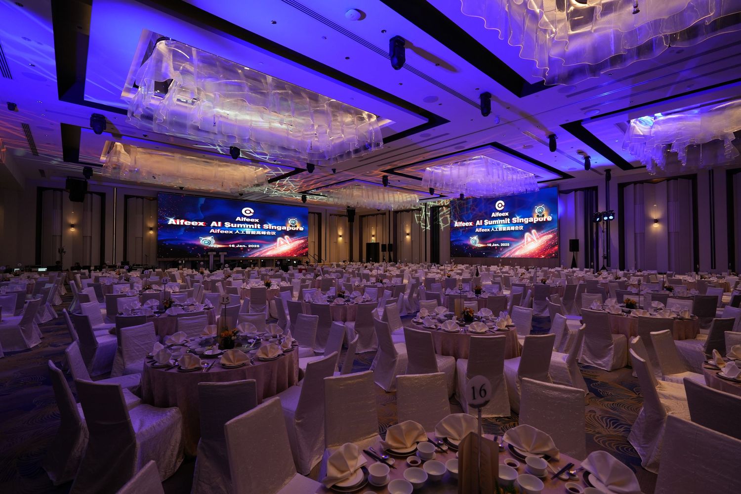 Elegant banquet hall with round tables, white chair covers, purple lighting, and large screens displaying a corporate event presentation.