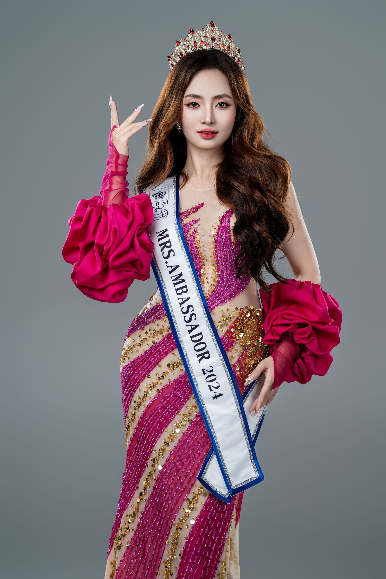Woman in pink sequin gown wearing Mrs. Ambassador 2024 sash and crown poses gracefully against gray background.