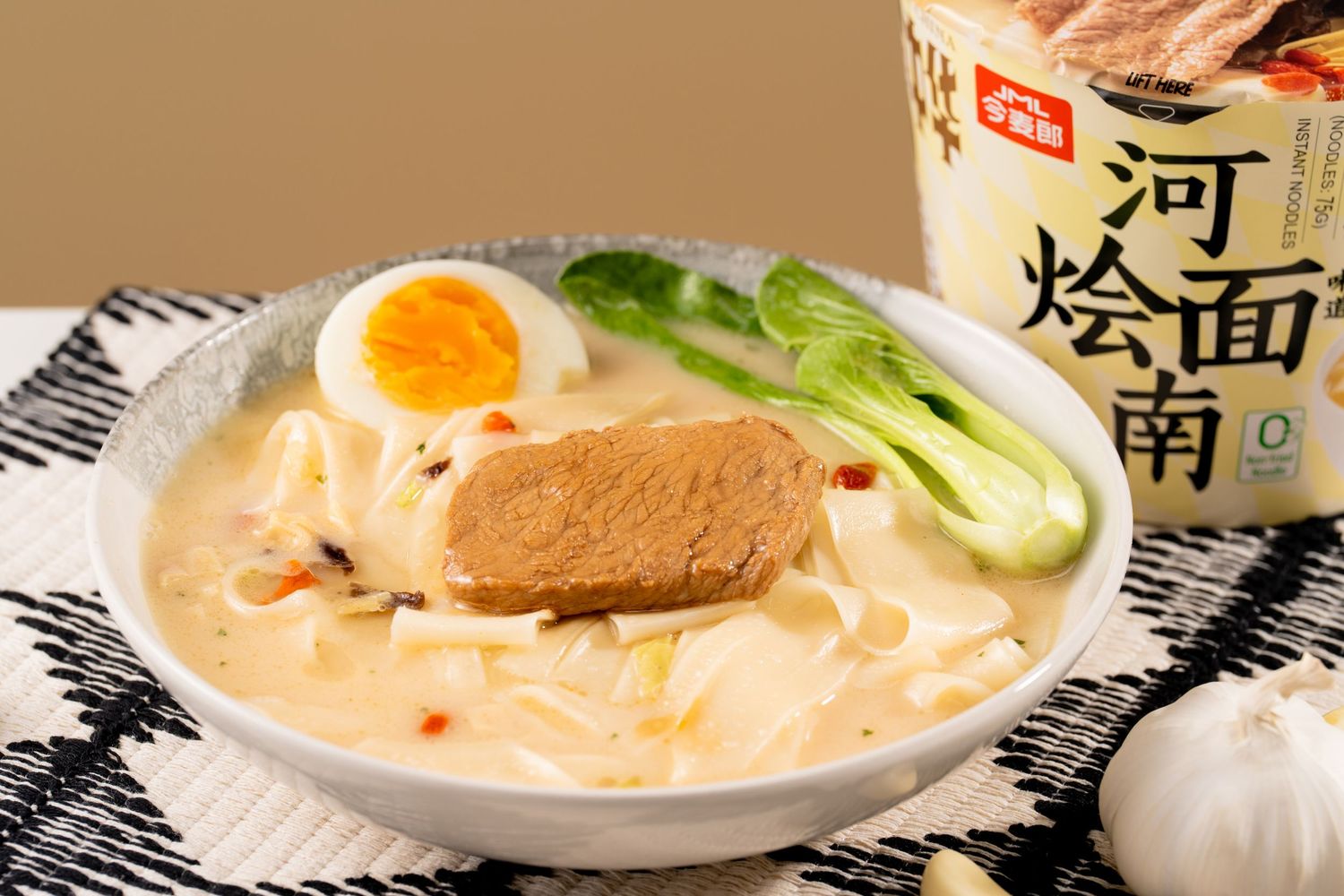 A bowl of creamy ramen with a boiled egg, bok choy, and meat slice, served next to a branded instant noodle cup.