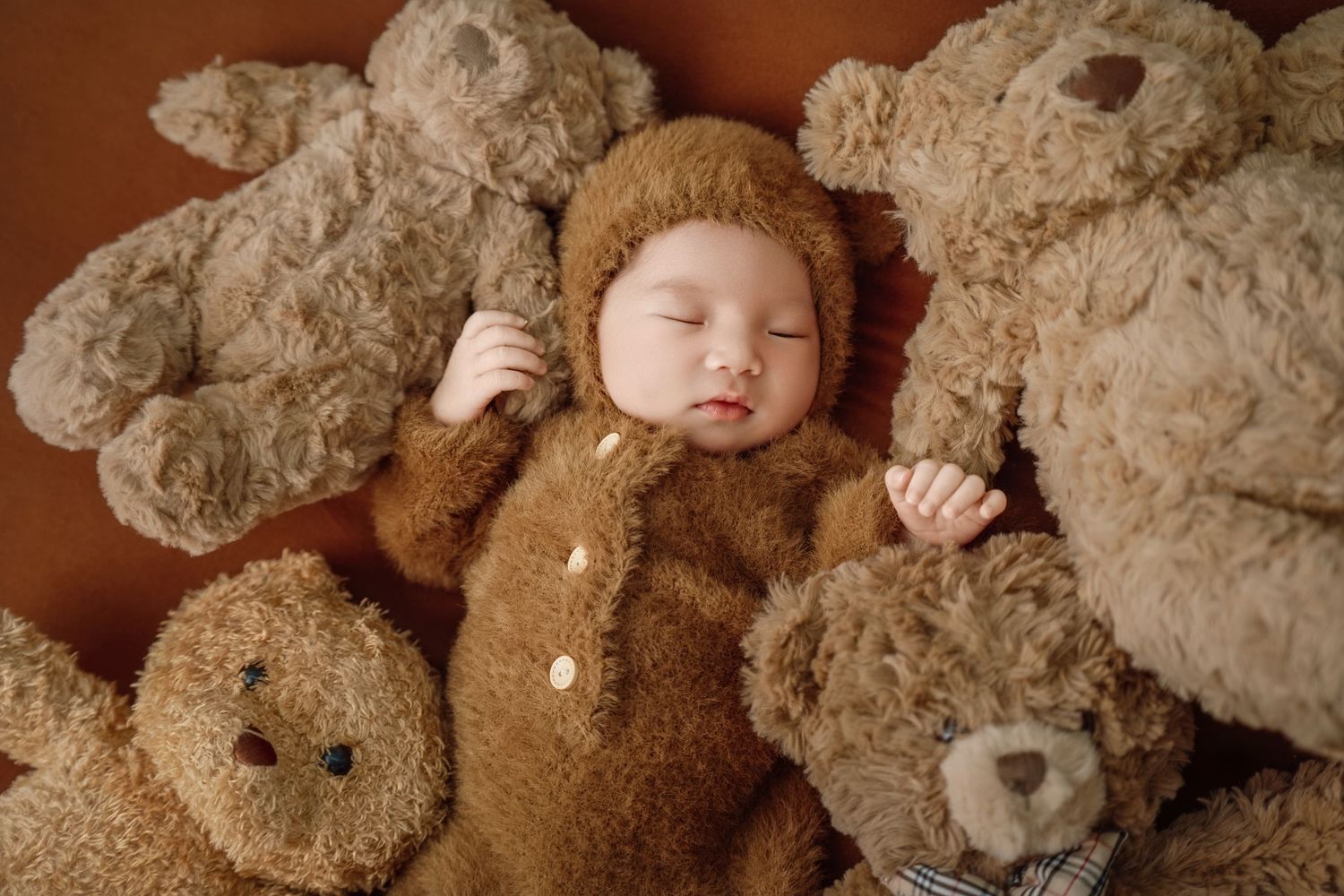 A sleeping baby in a brown teddy bear outfit surrounded by plush teddy bears on a warm brown background.