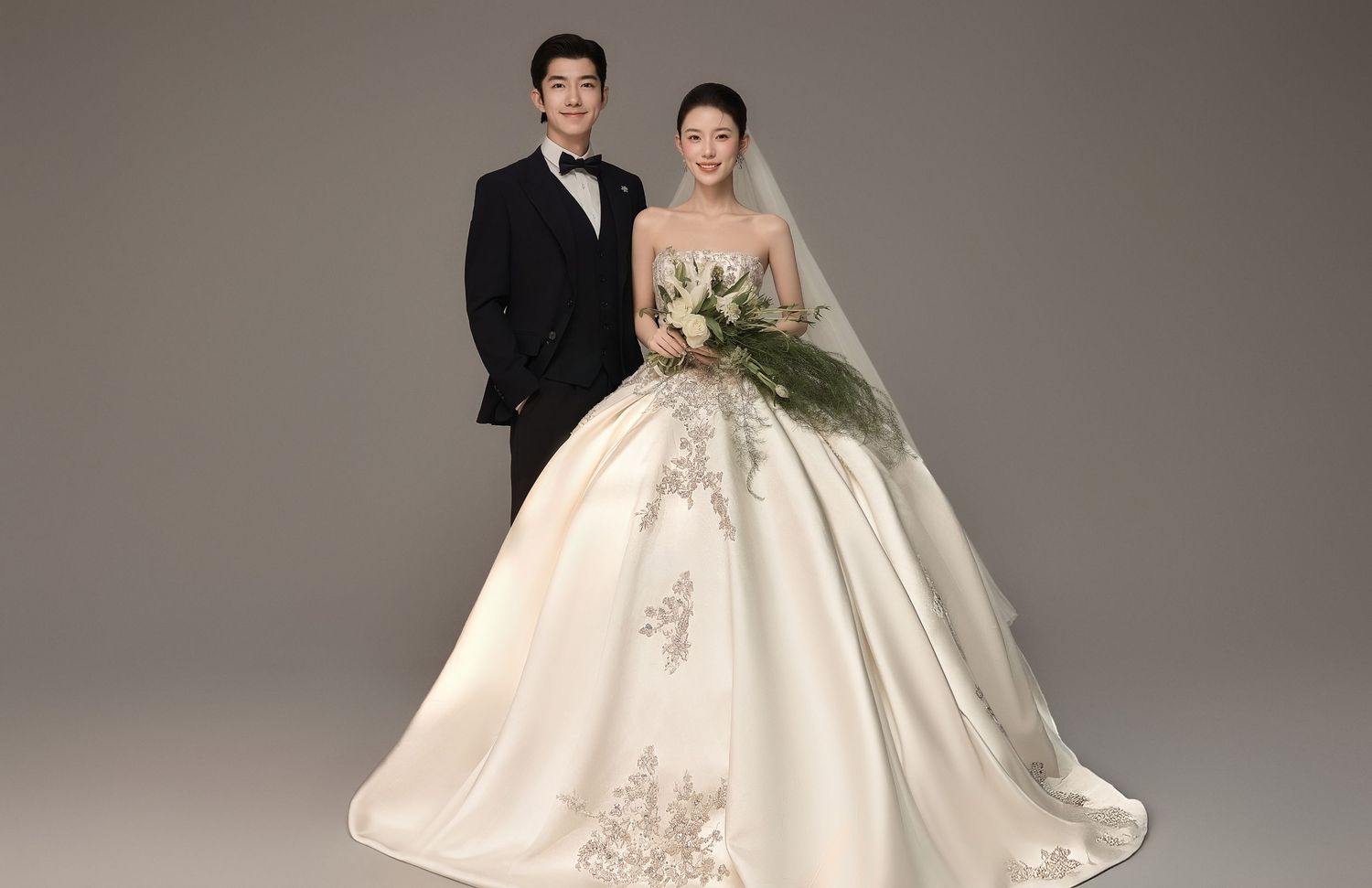 Elegant couple posing in wedding attire, bride in embroidered ball gown with veil holding bouquet, groom in black tuxedo.