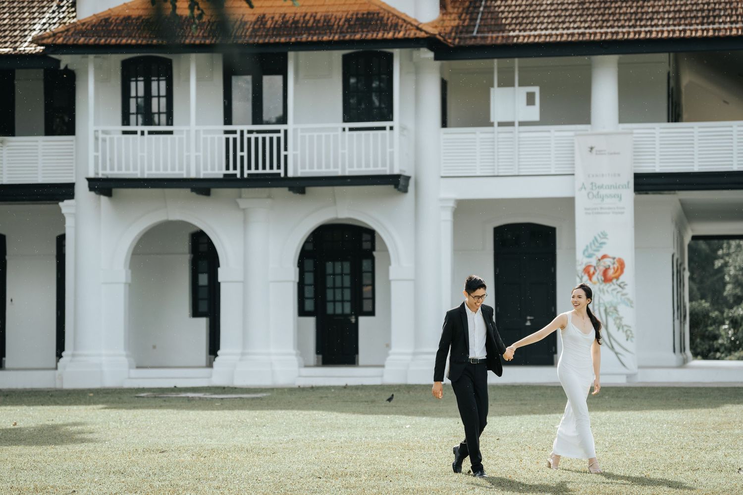 Wedding couple walks across a green lawn in front of a white colonial style building during their photo session.