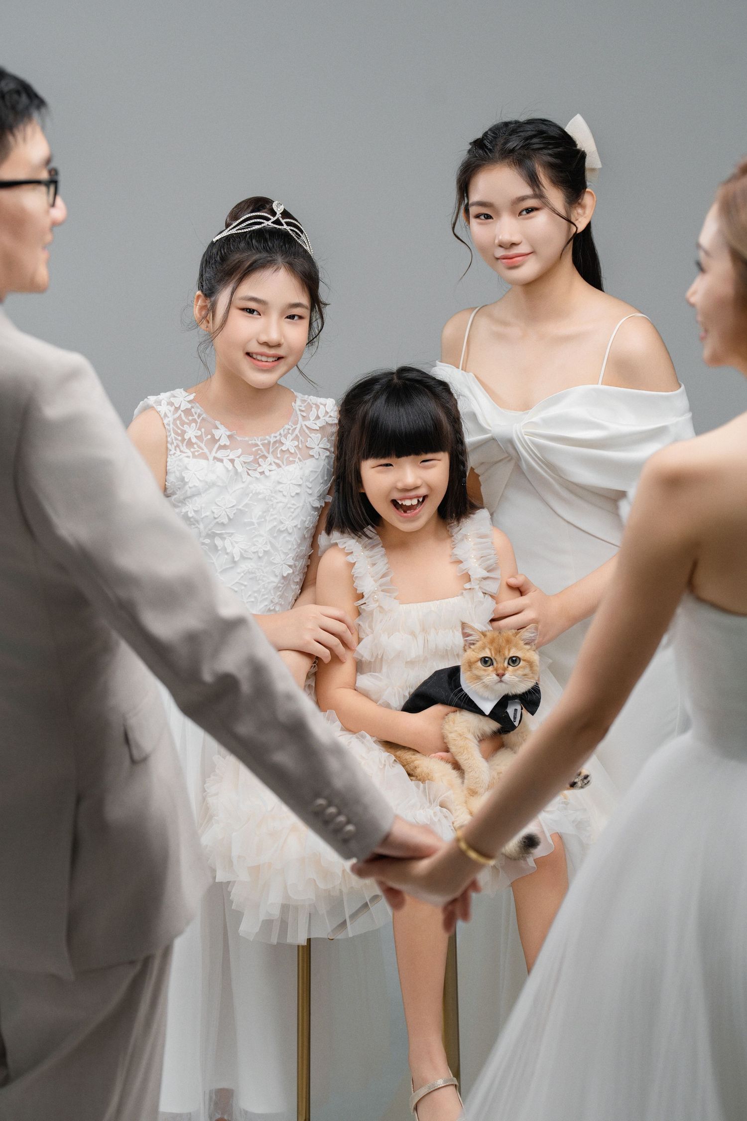 Asian family in white formal attire posing with a cat dressed in a tuxedo during a wedding photo shoot.