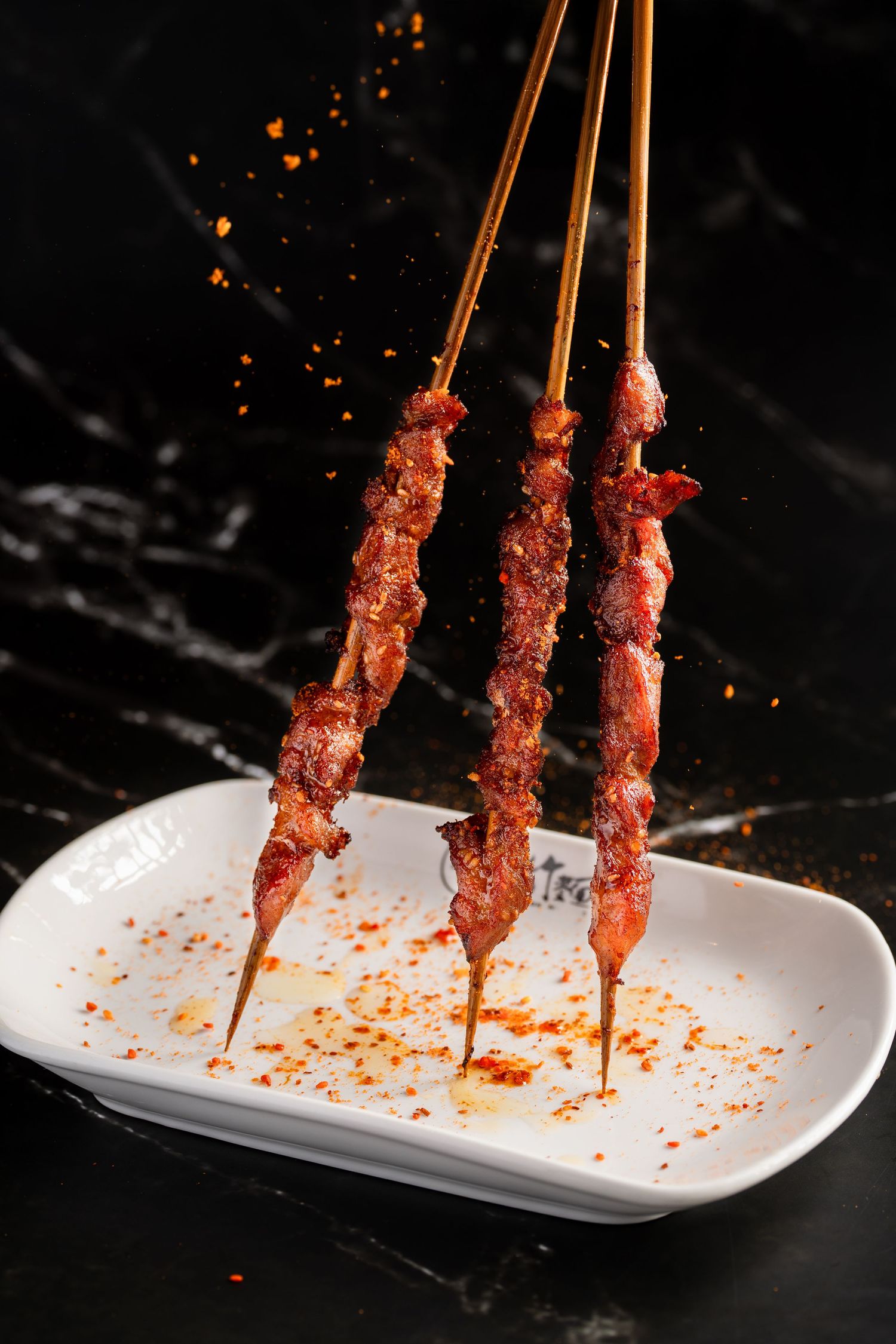 Three spicy meat skewers on wooden sticks served on a white plate with chili seasoning on dark marble background.