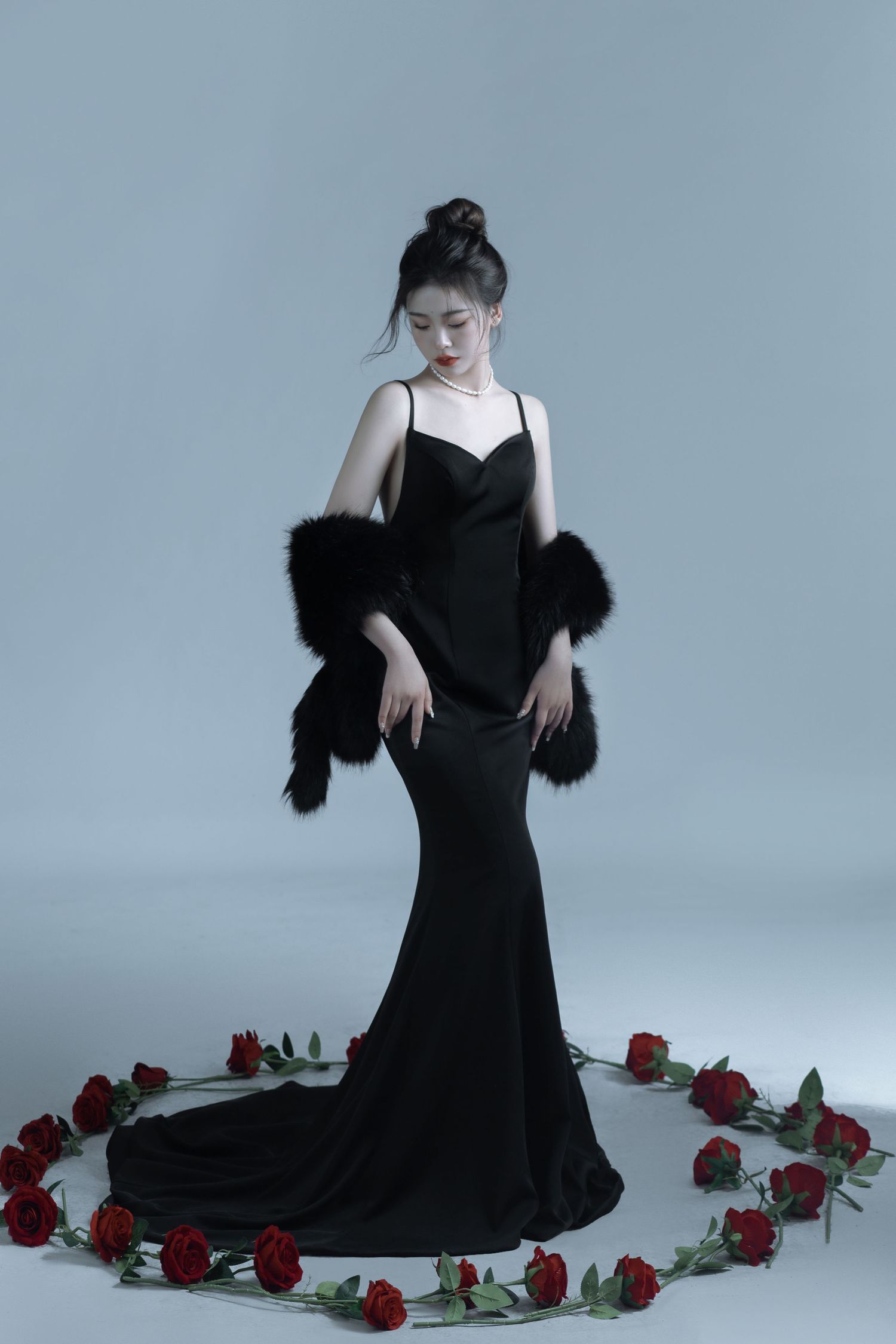 A dramatic fashion photoshoot with a black evening gown surrounded by scattered red roses against a light gray backdrop.