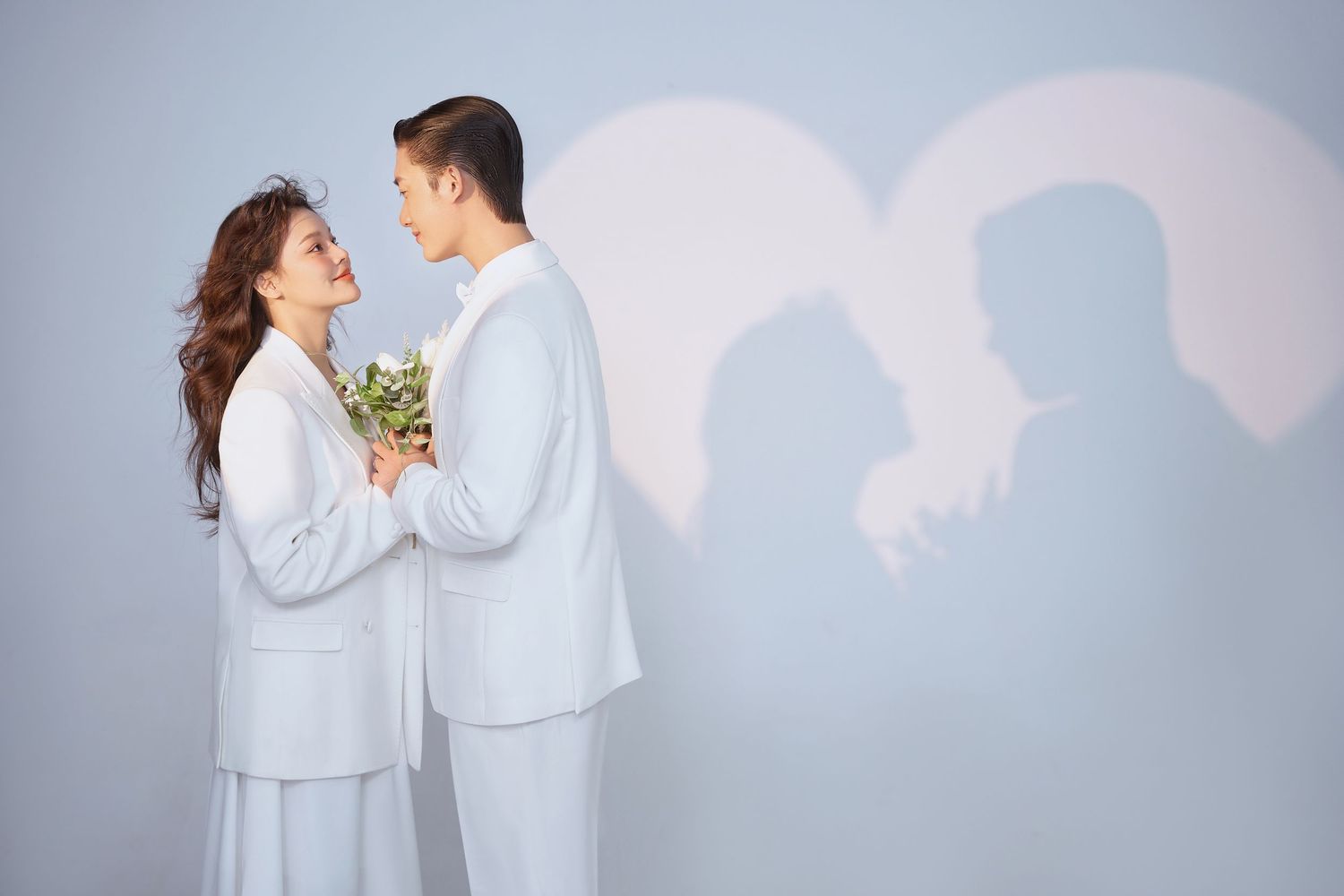 A romantic couple in matching white outfits stands against a gray wall with heart shadow silhouettes behind them.