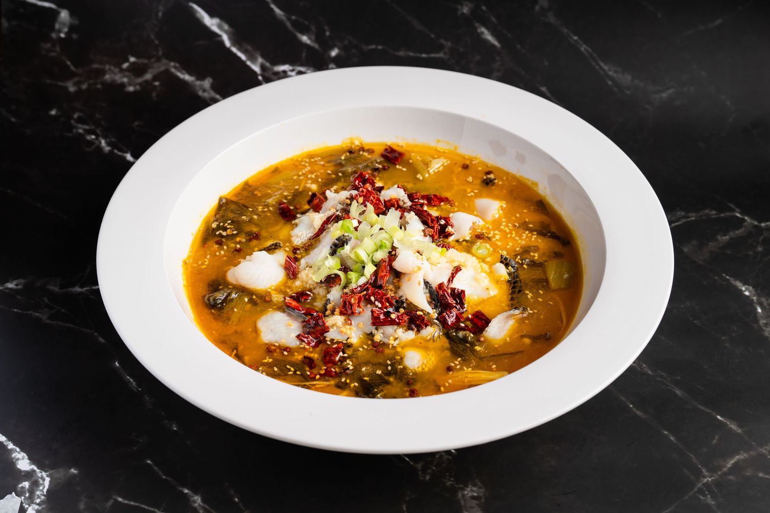 Spicy Chinese fish fillet soup in white bowl with chili peppers, scallions, and rich broth on marble surface.
