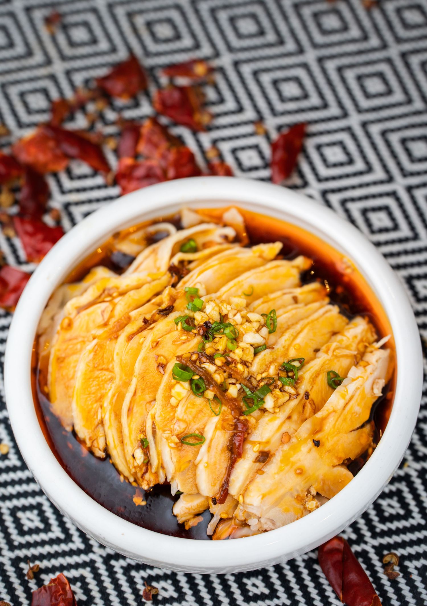 Sliced poached chicken in spicy chili oil sauce topped with crushed peanuts and green onions in a white bowl.