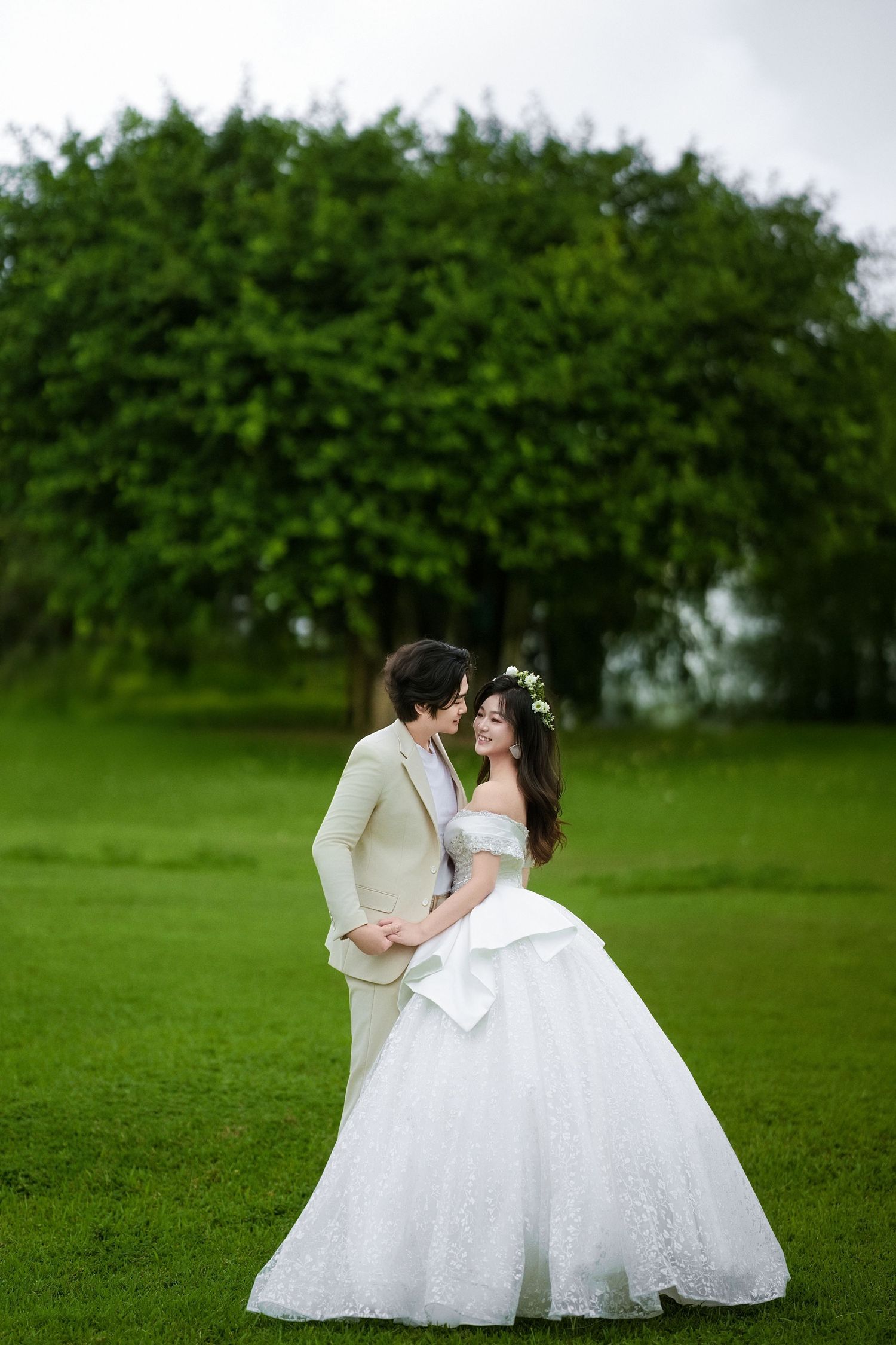 A couple in wedding attire shares romantic moments on a green lawn with lush trees in the background.