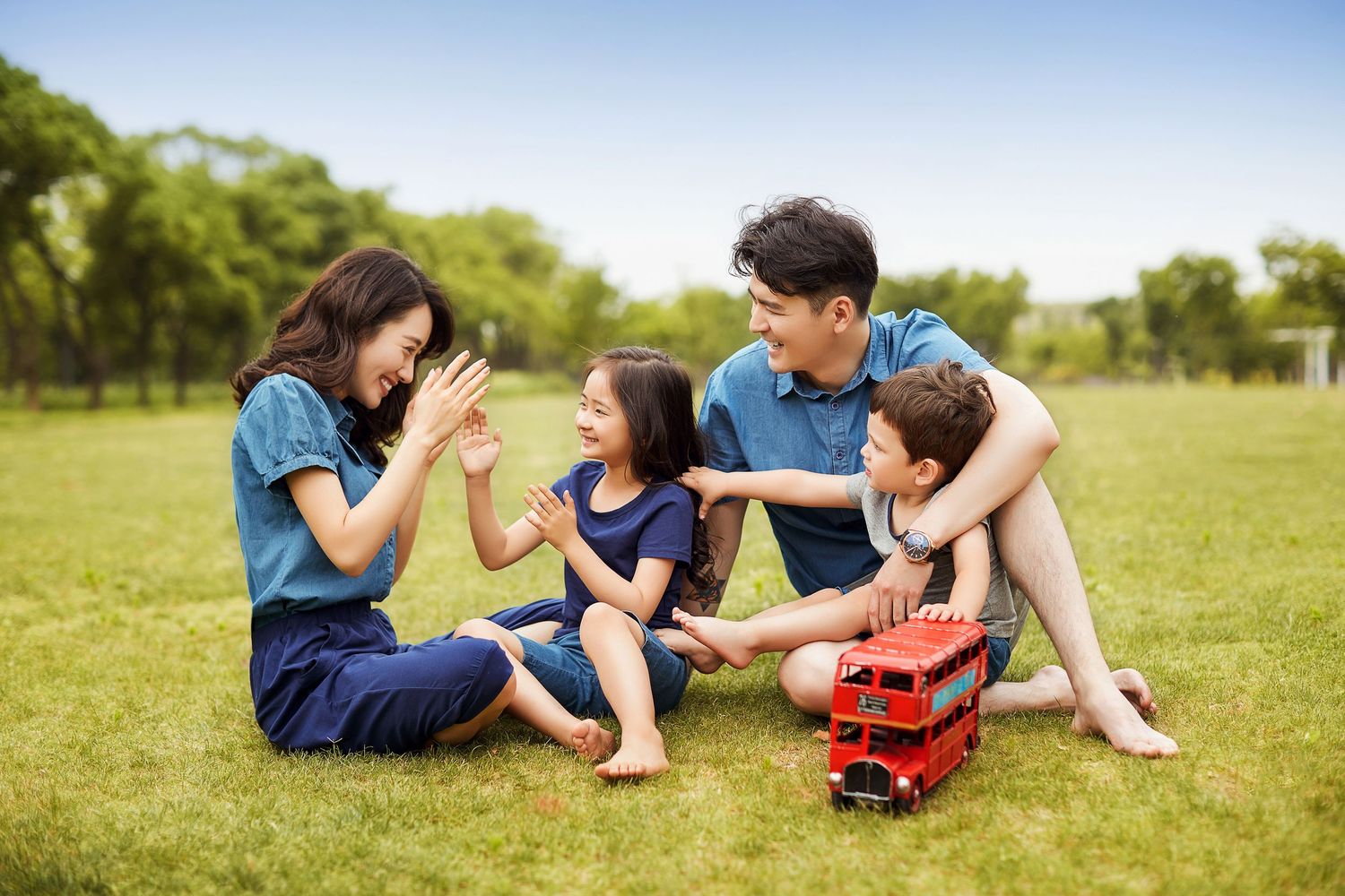 Asian family of four sitting on green grass in a park, playing and laughing together on a sunny day.