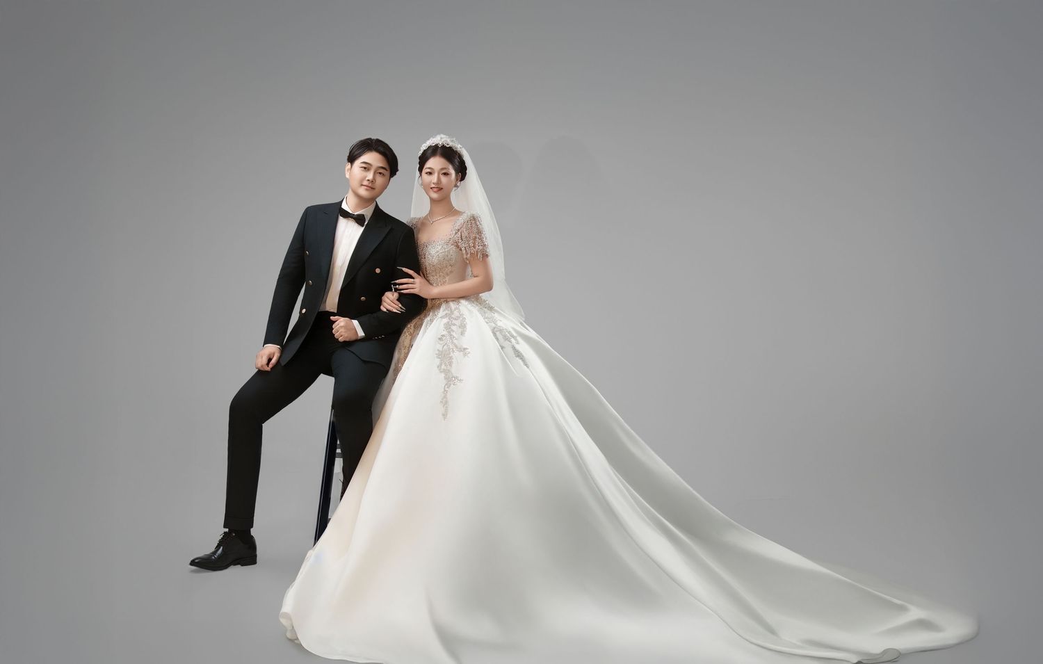 A couple poses for a Korean wedding photo, the groom in a black tuxedo and bride in an elegant white ball gown.