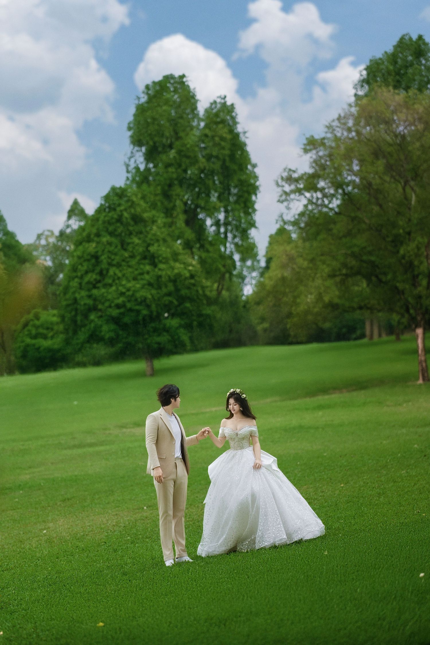 A bride in a white ball gown and groom in beige suit hold hands on a lush green lawn with trees and blue sky.