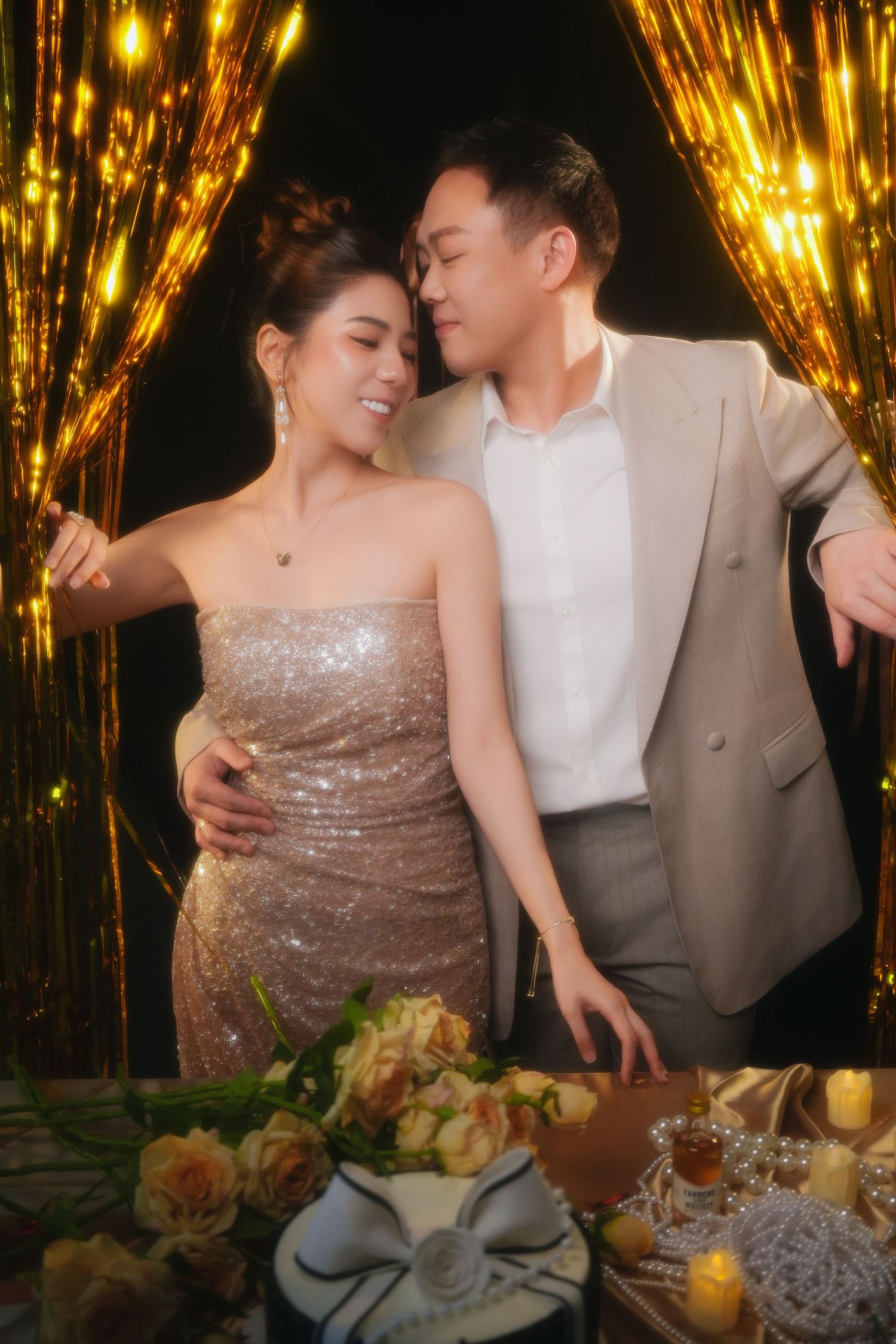 A romantic couple poses at an elegant dinner party with sparkling gold decorations and floral arrangements on the table.