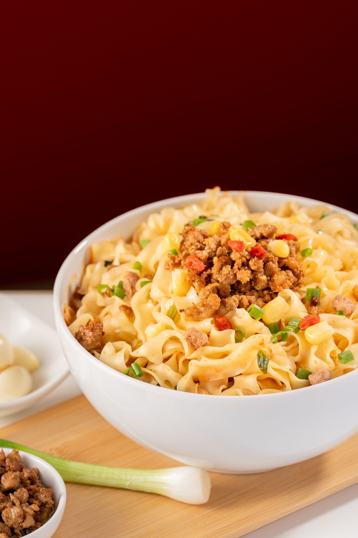 A white bowl of noodles topped with seasoned ground meat, red peppers, and green onions on a wooden board.