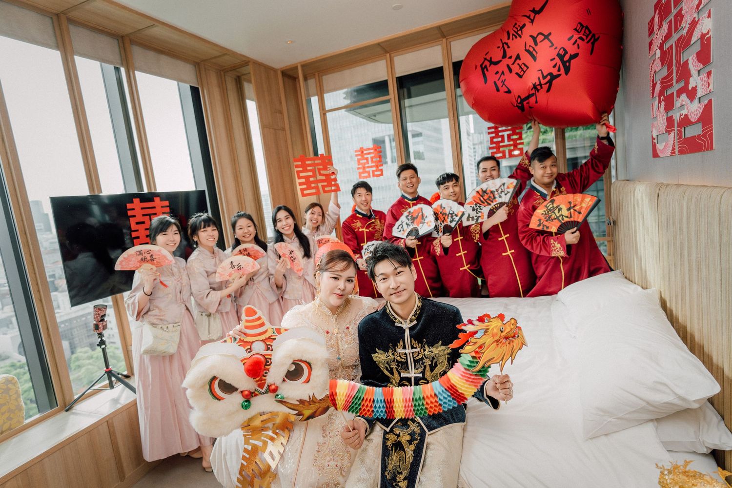 A joyful Chinese wedding party in traditional red attire celebrates in a modern hotel room with lion dance props and decorations.