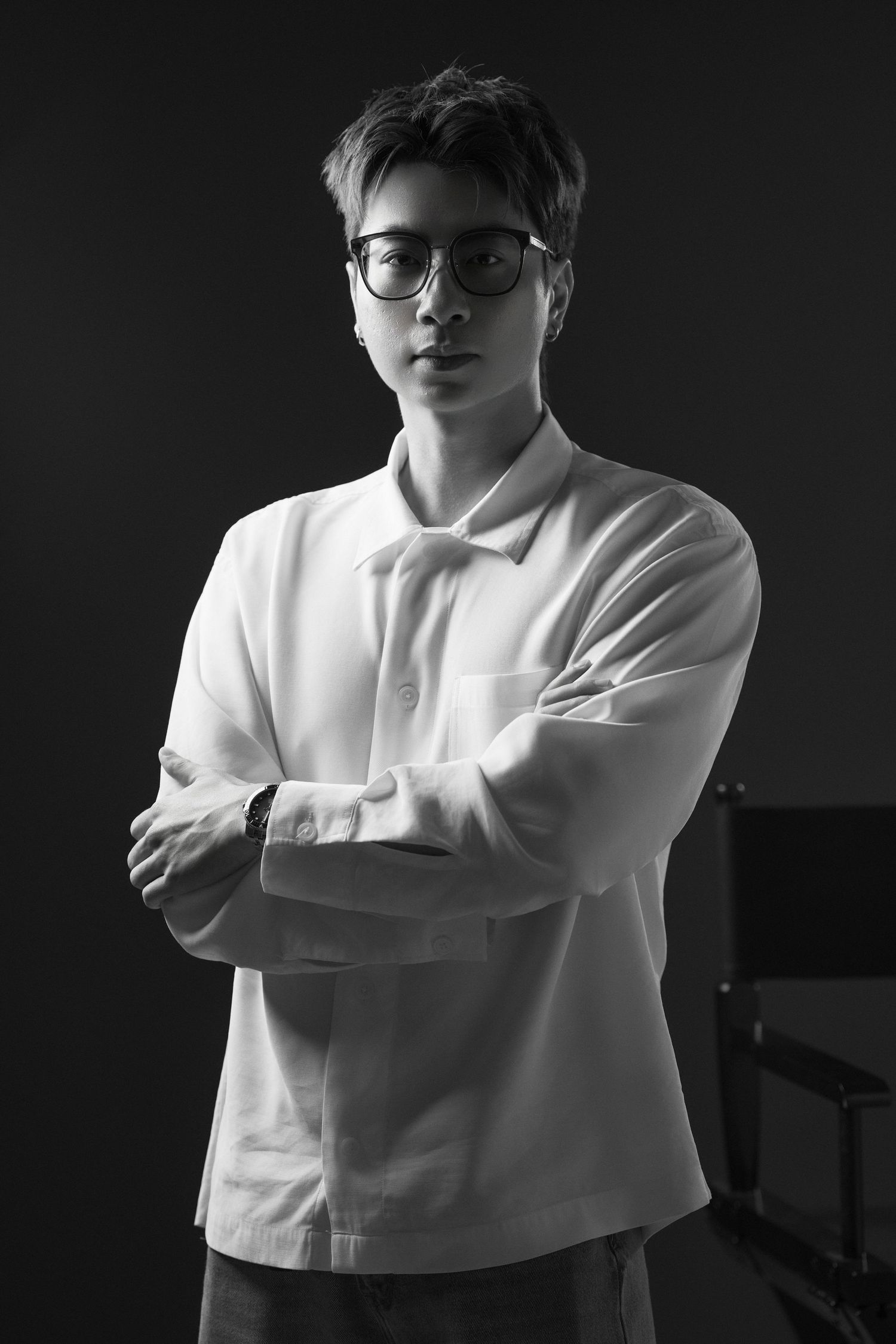 Black and white portrait of a young man in white shirt, arms crossed, wearing glasses, studio background.