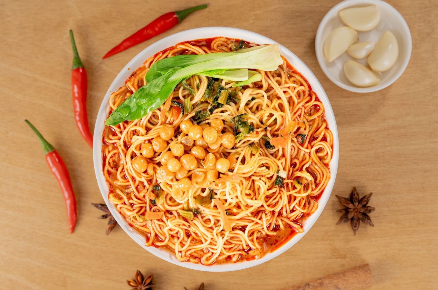 Spicy noodle bowl with chickpeas, chili peppers, garlic, star anise, and fresh herbs on a wooden surface.