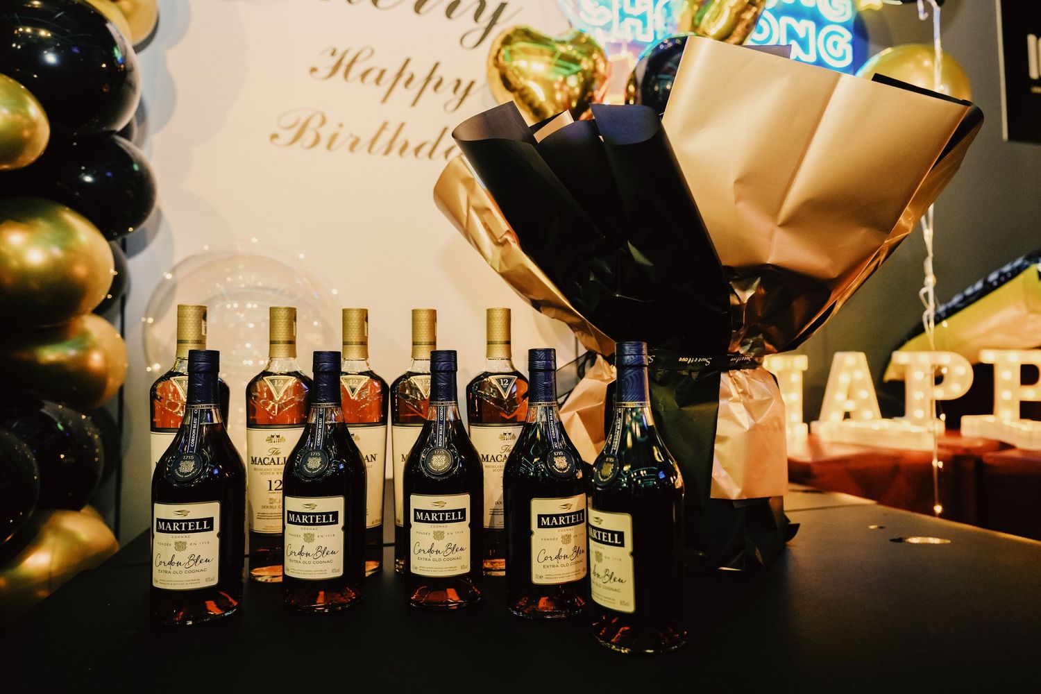 Martell cognac bottles displayed at a birthday party with gold and black balloons and Happy Birthday backdrop.