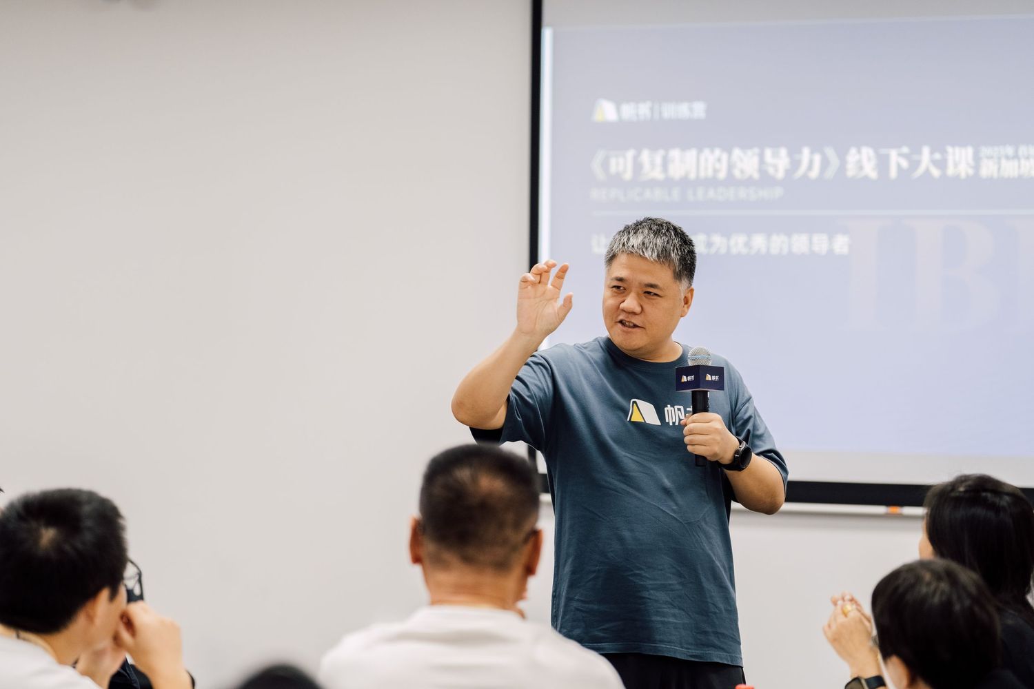A man in a blue t-shirt gestures while speaking at a seminar in front of a presentation screen with Chinese text.