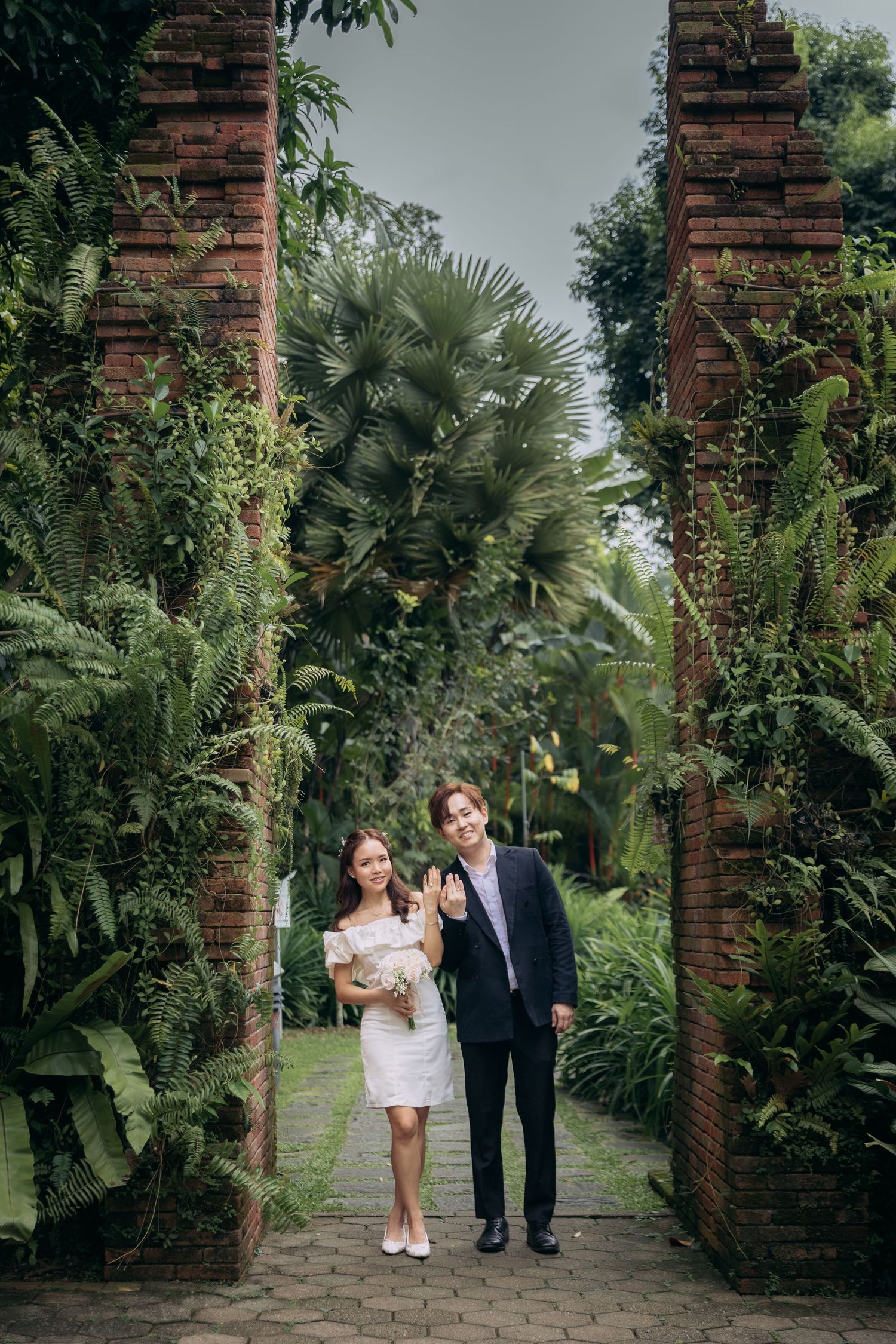 A couple strolls along a tropical garden path lined with brick pillars and lush palm trees under natural lighting.