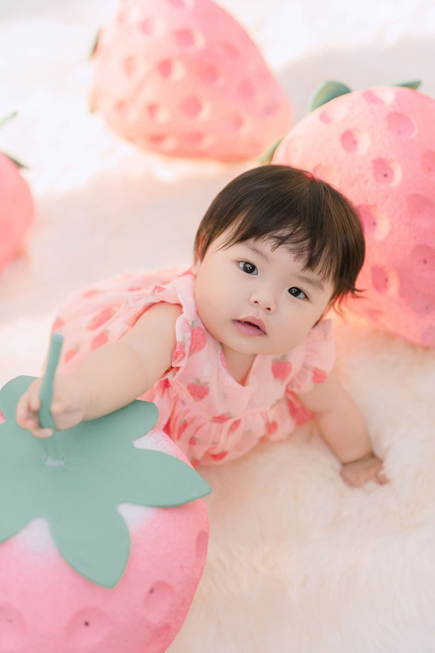 A cute baby girl in a pink strawberry dress poses among large decorative strawberry props on a fluffy white rug.